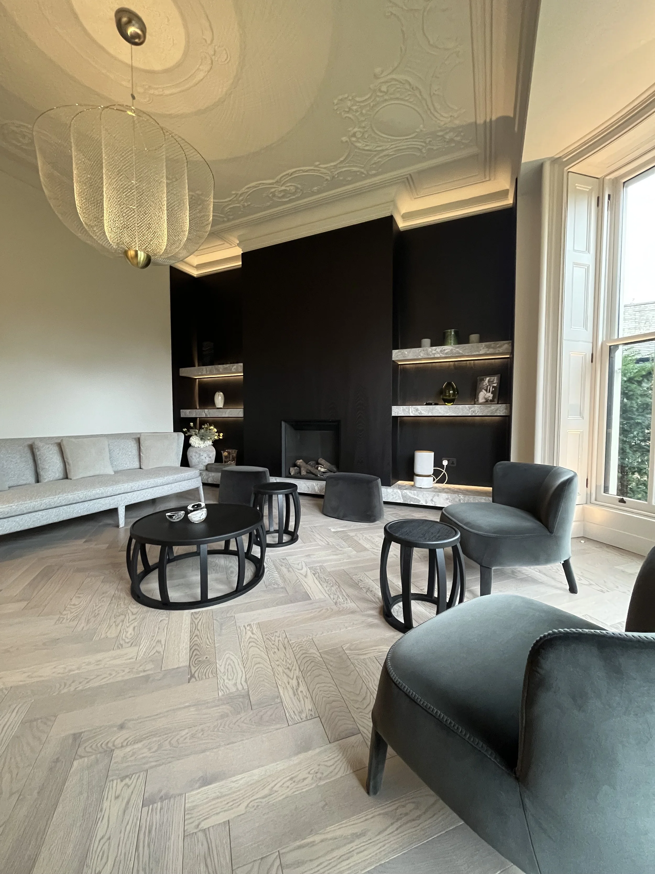 A modern living room with a beige sofa, black chairs, small coffee tables, a fireplace with a black wall behind it, open shelving with decorative items, a large window with wooden shutters, a light fixture hanging from the ceiling, and ornate ceiling