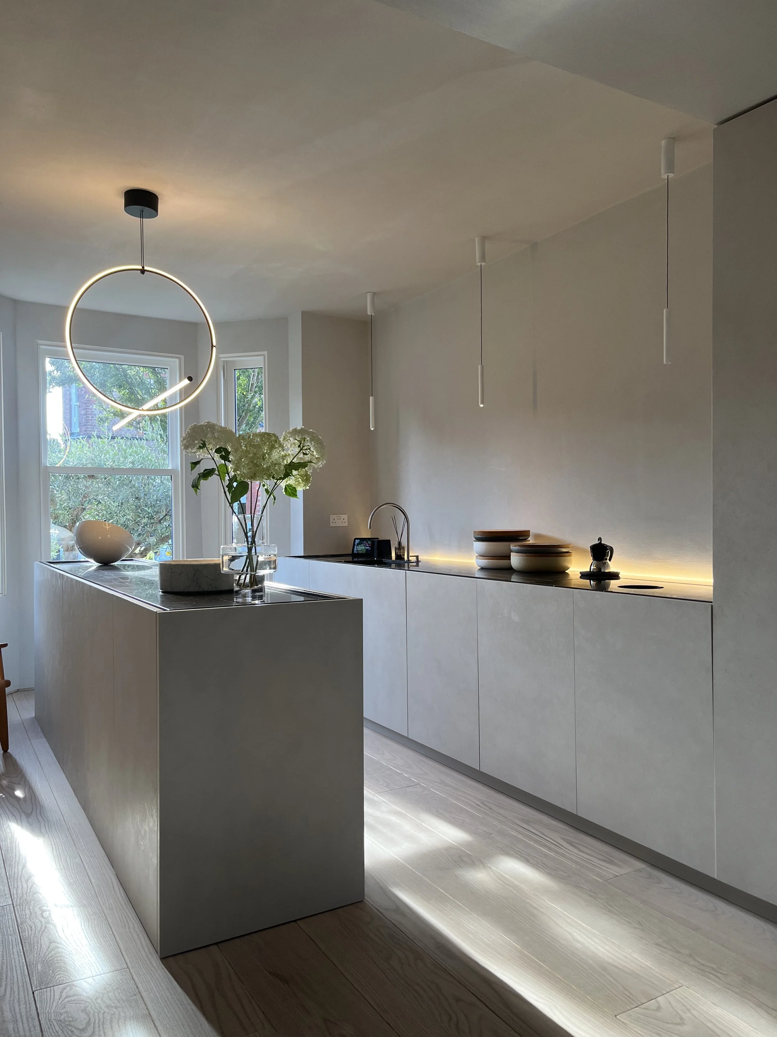 Modern kitchen with white cabinetry, a white island with a flower vase, objects on the countertop, and pendant lighting, with large windows showing outdoors.