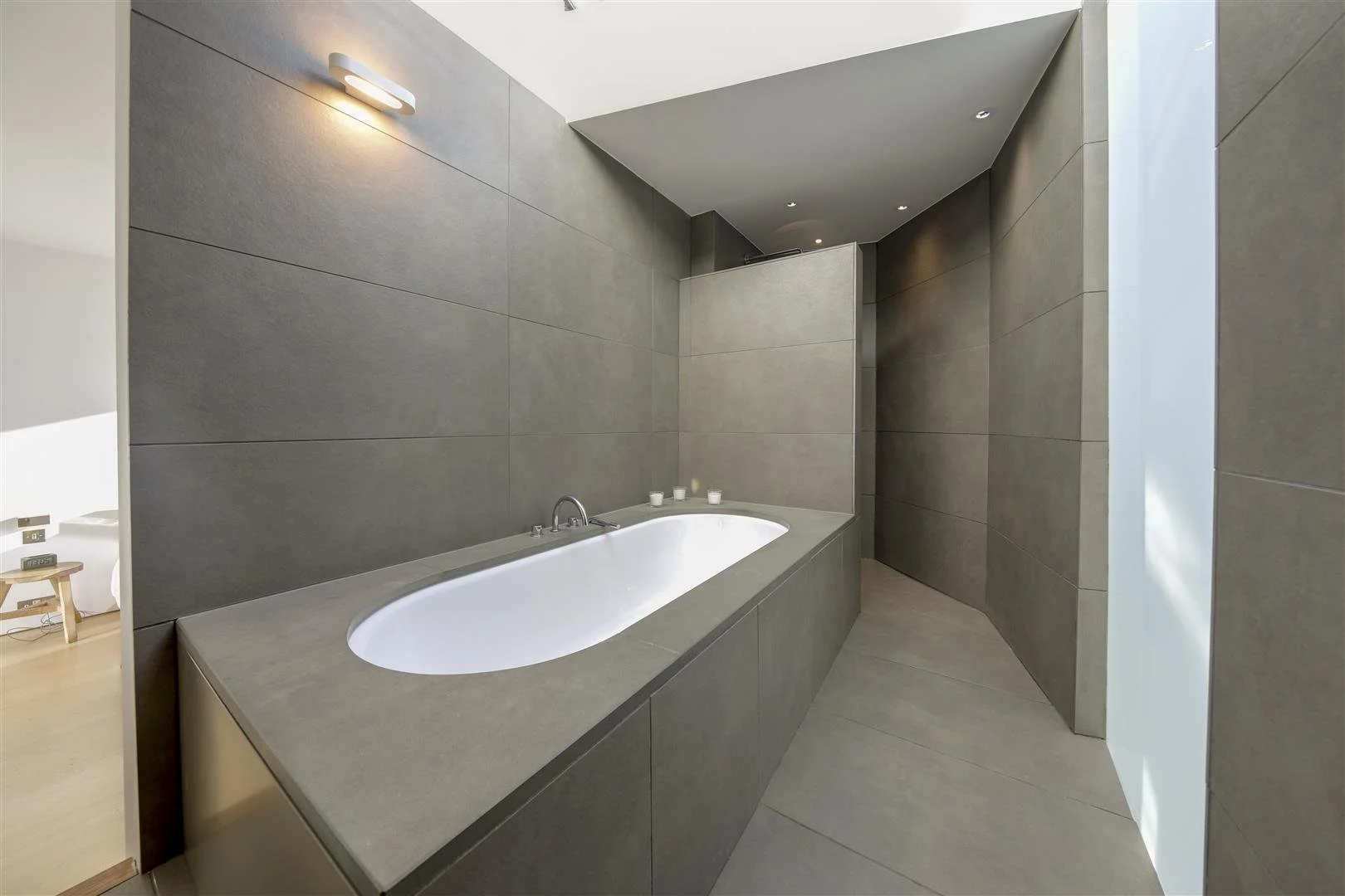 Modern bathroom with a built-in bathtub surrounded by tiled walls in neutral gray tones, with four small candles on the edge of the tub, and soft lighting.
