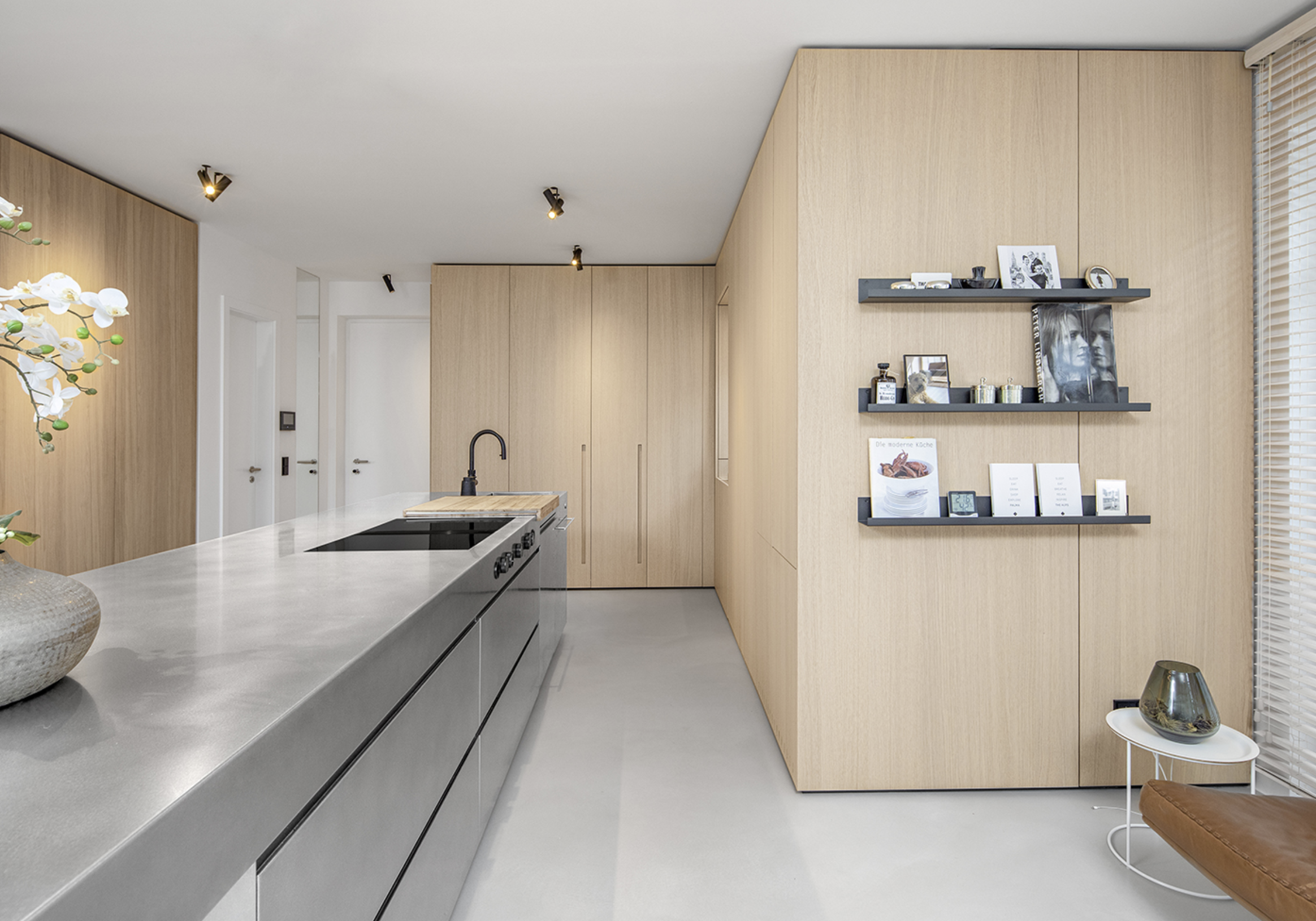 Modern kitchen with light wood cabinetry, a white countertop with a built-in black electric cooktop, a black kitchen faucet, and minimalist decor including books, framed photos, and small decorative items on black shelves on a wood-paneled wall.