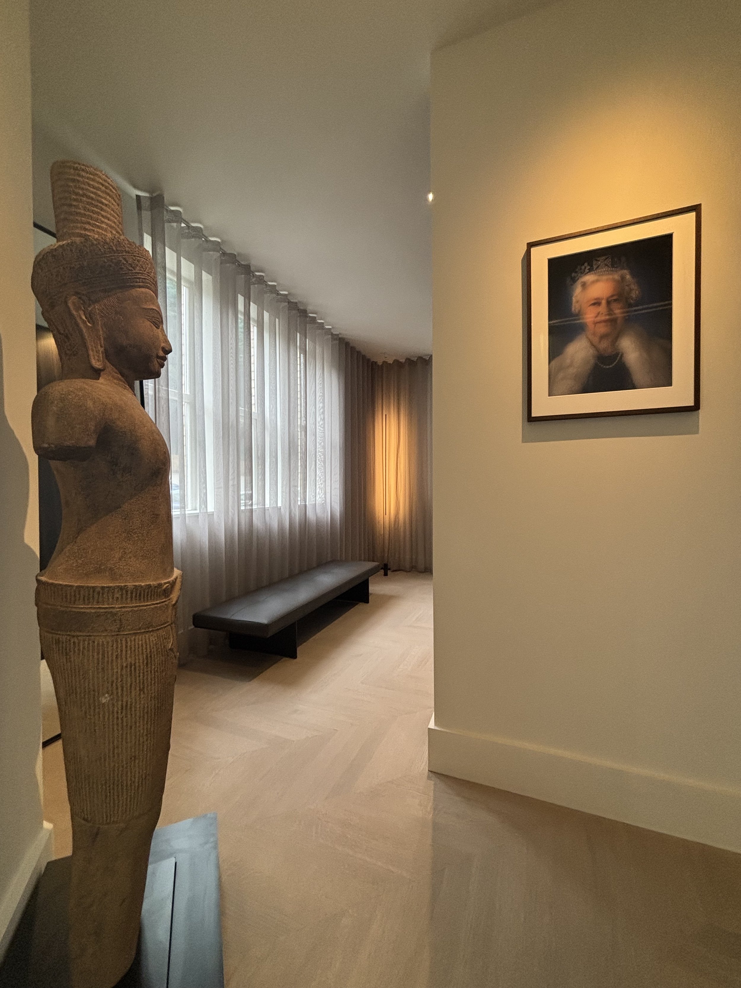Interior of a room with a stone sculpture of a woman on the left, a framed photograph of Queen Elizabeth II on the right wall, a bench against the window, and beige curtains with natural light.