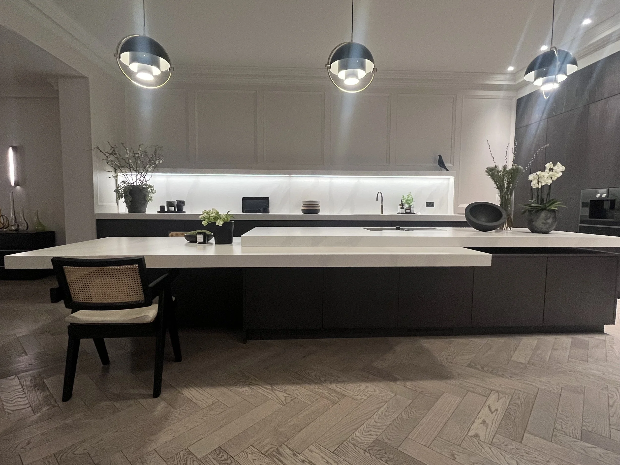 Modern kitchen with white countertops, black cabinets, pendant lights, and decorative plants.