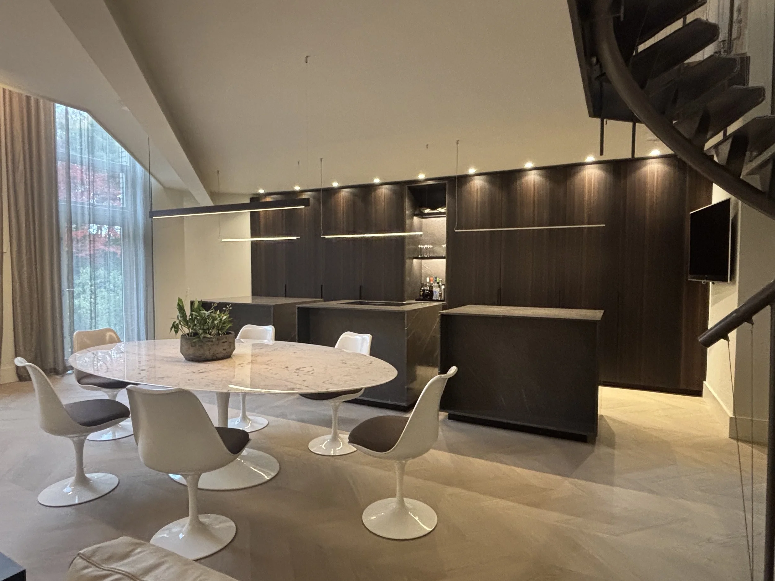 Modern kitchen and dining area with a round marble table and six white and gray chairs. Dark wooden cabinets, a small bar area, and a wall-mounted TV. Large windows with sheer curtains and a staircase on the right.