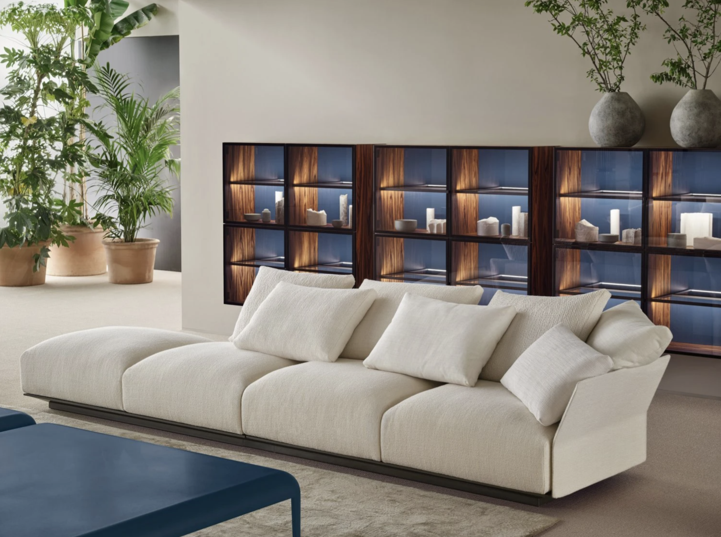 Modern living room with a white sectional sofa, a blue coffee table, and wooden display cabinets with shelves of decorative objects and candles, complemented by large potted plants and minimalist decor.