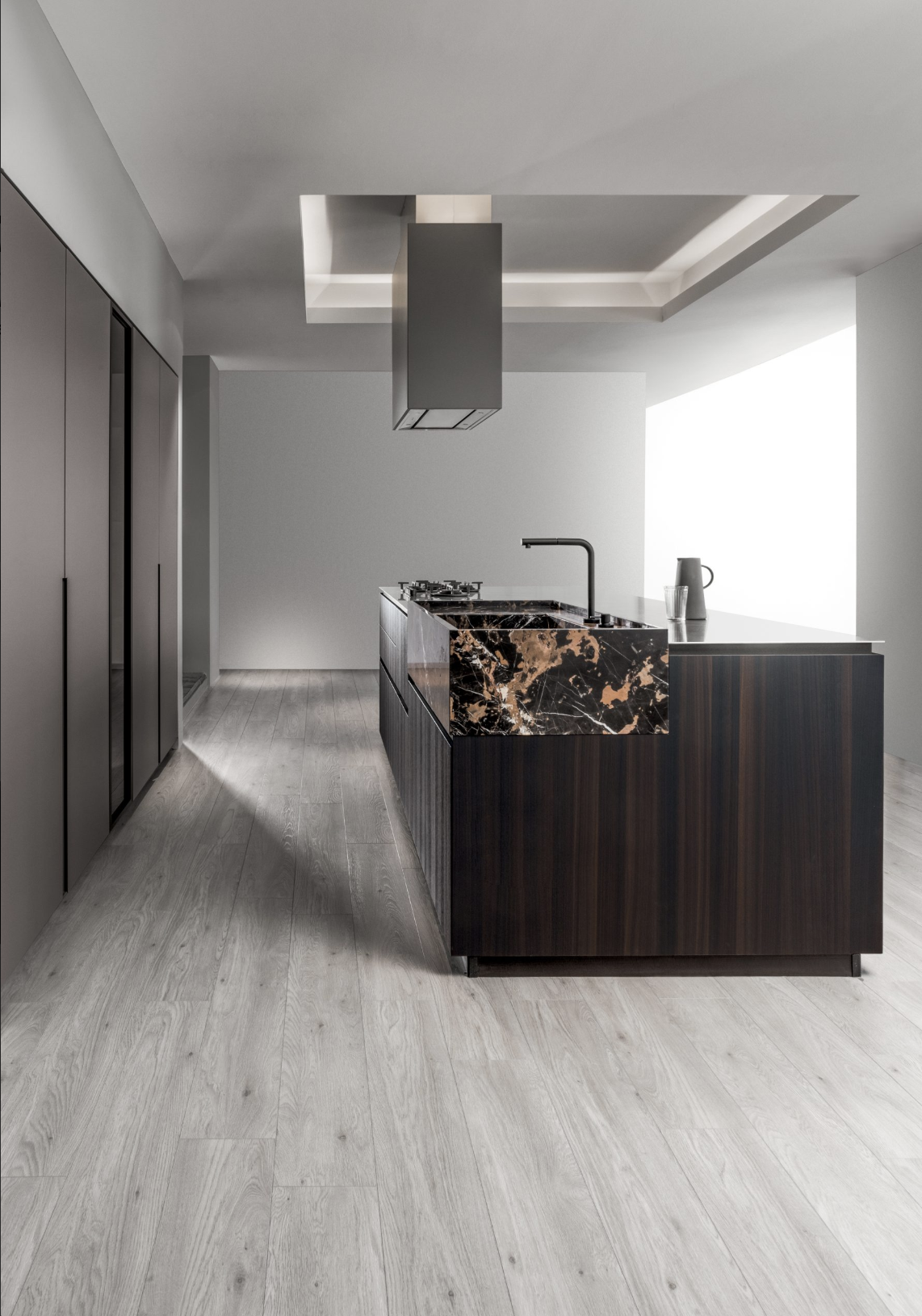Modern kitchen with a black marble island and sink, gray cabinets on the left, light wood flooring, and a minimalist ceiling with recessed lighting.