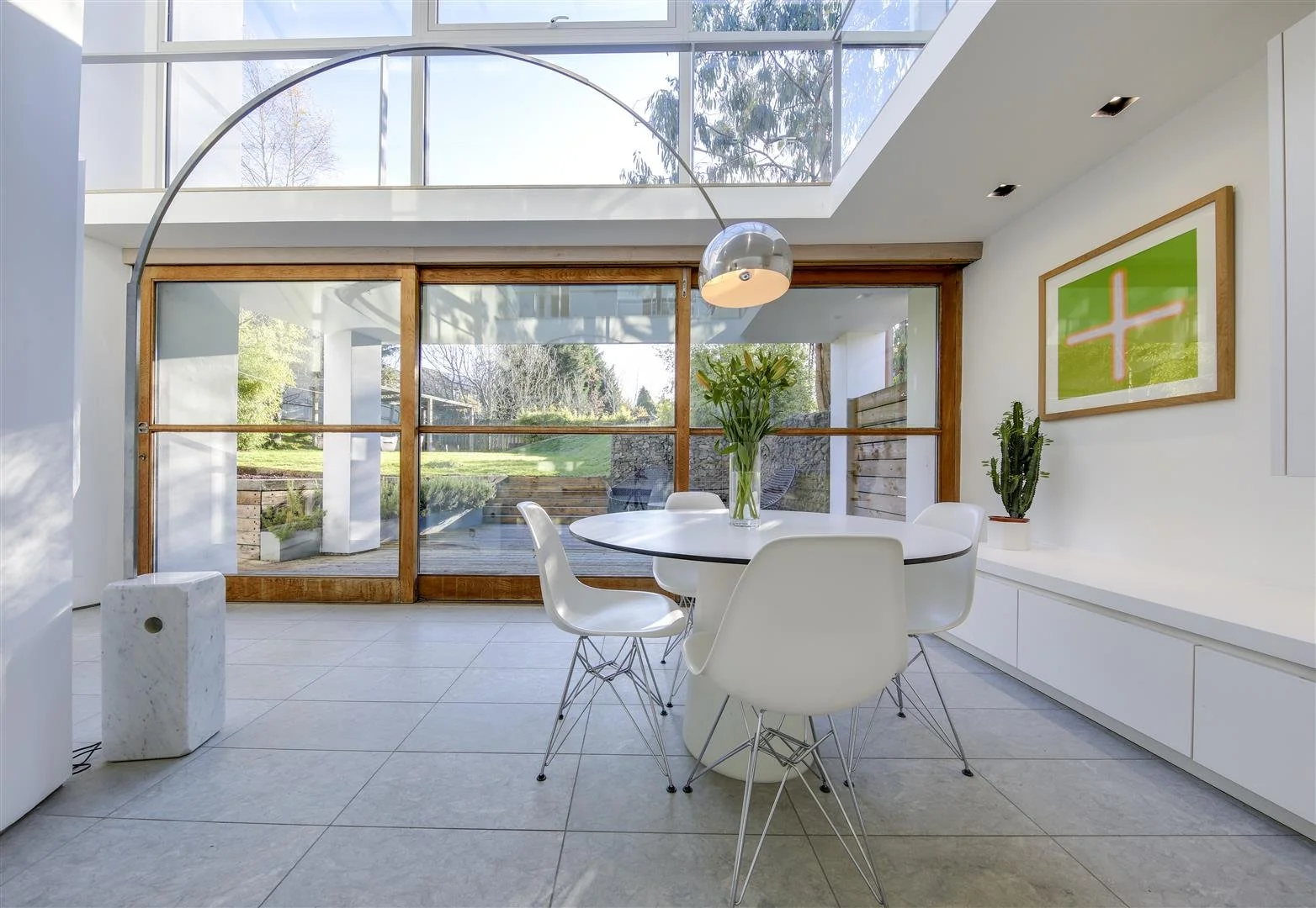 Modern dining area with a round white table, four white chairs, a vase of lilies, and a large window overlooking a garden