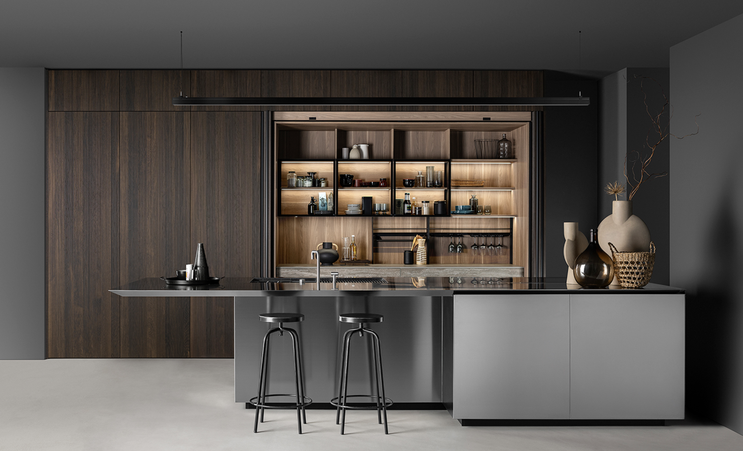 Modern kitchen with dark wood cabinets, gray island with black bar stools, open wooden shelves with various containers, and decorative vases with dried branches.