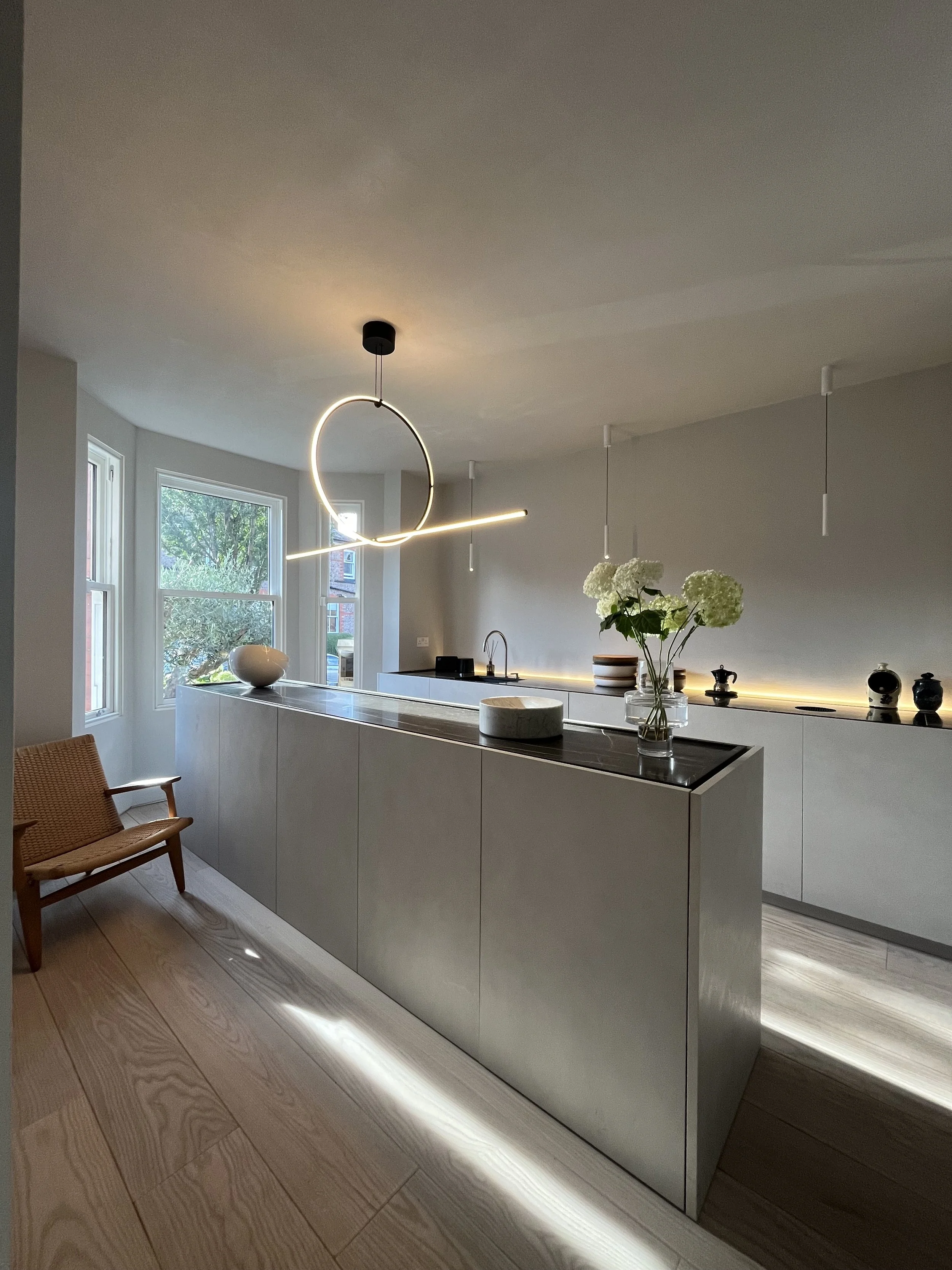 Modern kitchen with sleek white cabinets, a black countertop island, a vase with white flowers, and contemporary hanging light fixtures, adjacent to a sitting area with wooden furniture and large windows letting in natural light.