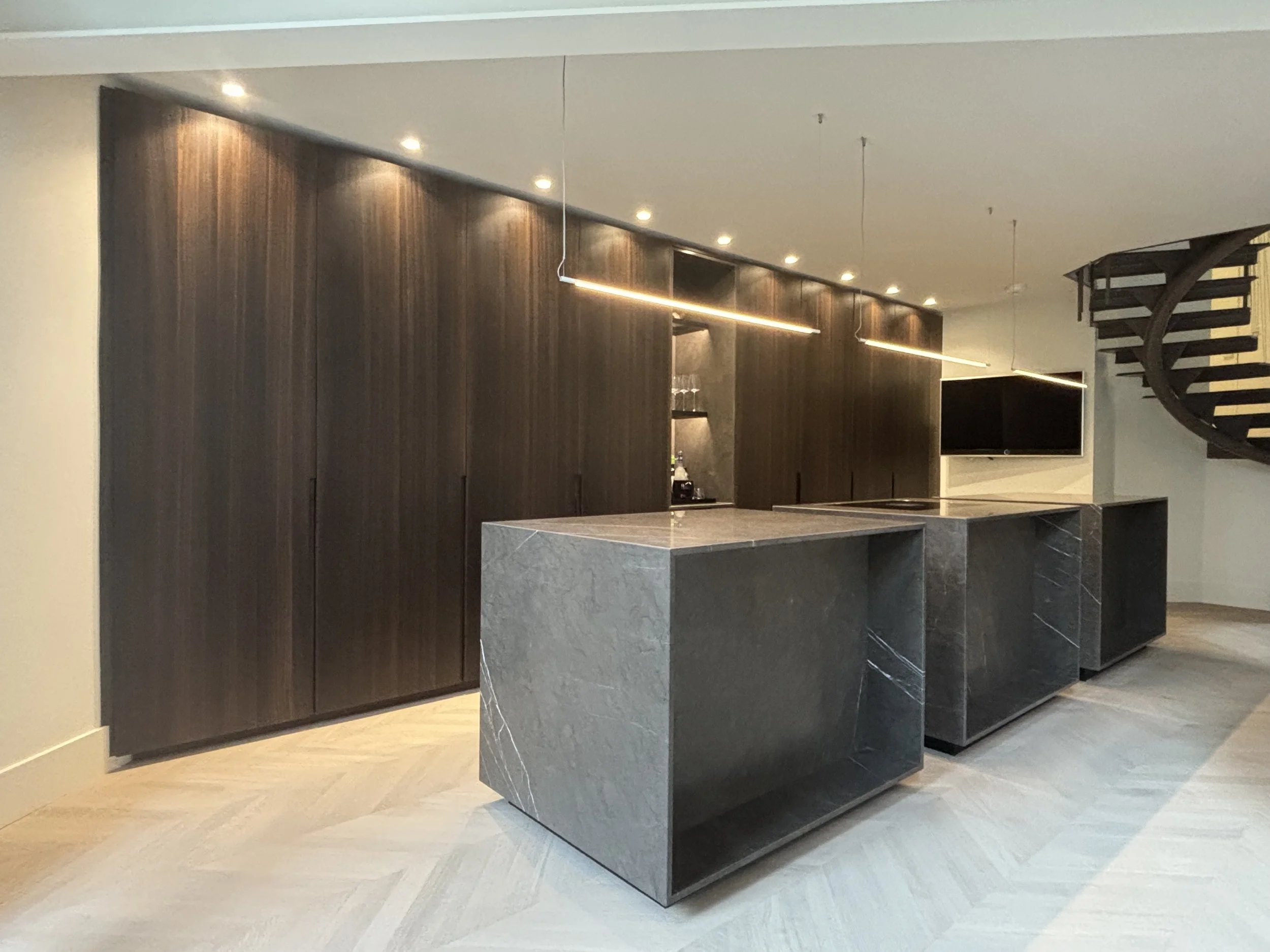 Modern kitchen with dark wood cabinetry, sleek gray marble island, and a flat-screen TV mounted on the wall. Spiral staircase visible on the right. Bright overhead lighting.