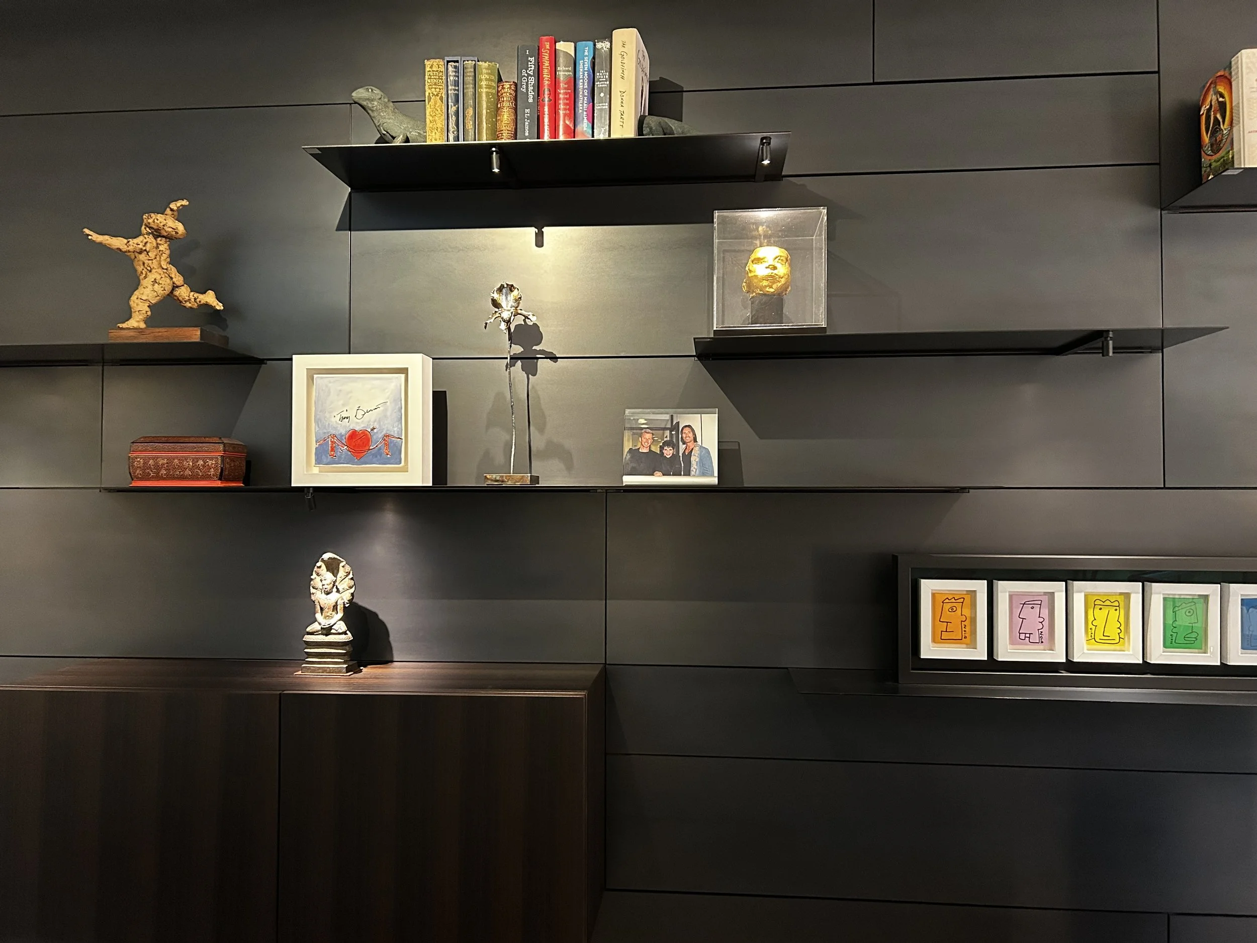 Wall-mounted shelves displaying various decorative objects, including books, sculptures, a framed photo, and colorful framed artwork, on a dark gray paneled wall.