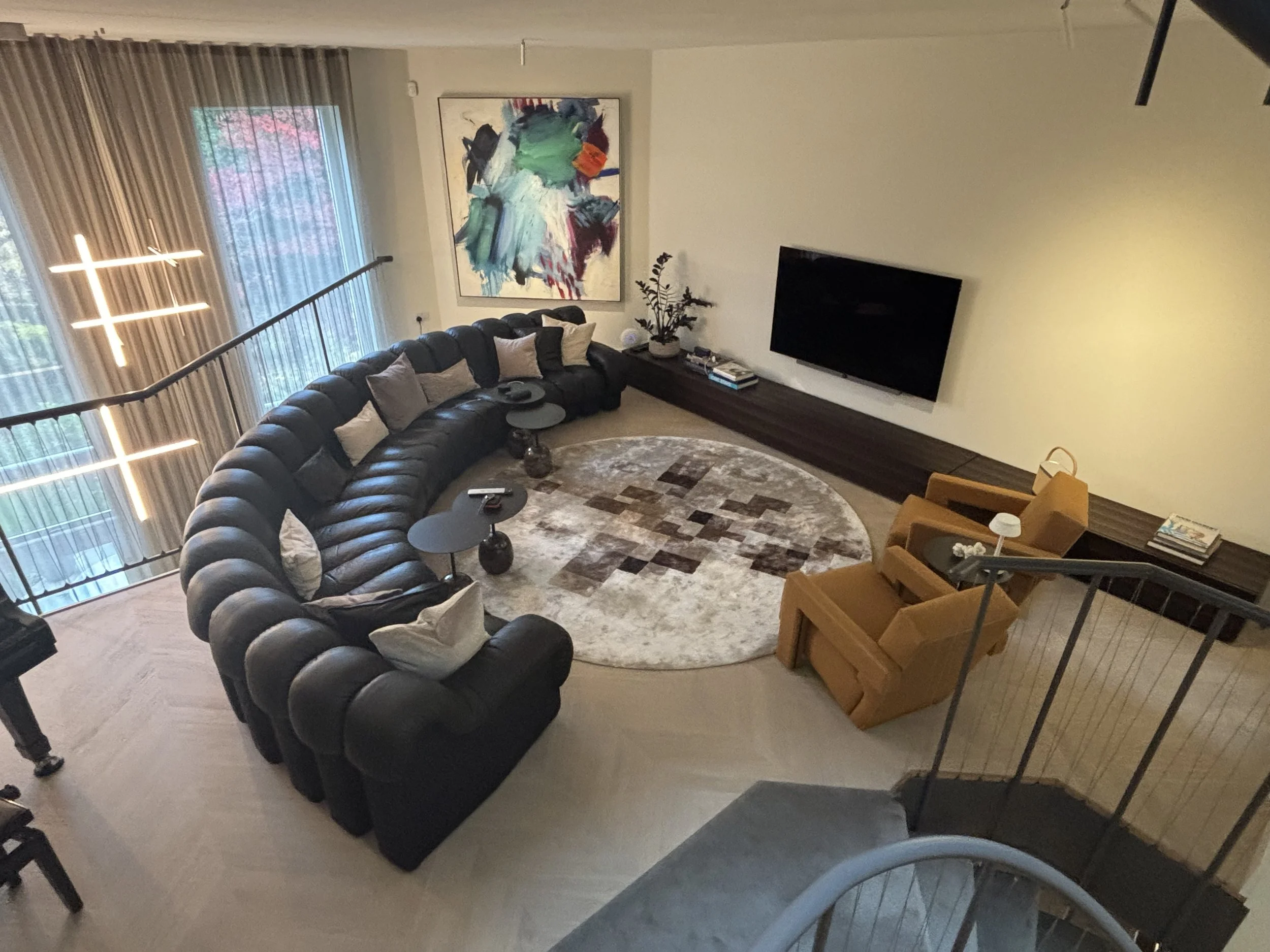 A high-angle view of a modern, well-decorated living room featuring a large black leather sectional sofa with pillows, two mustard-colored armchairs with pillows, a circular area rug with a geometric pattern, a long wooden media console, a wall-mount