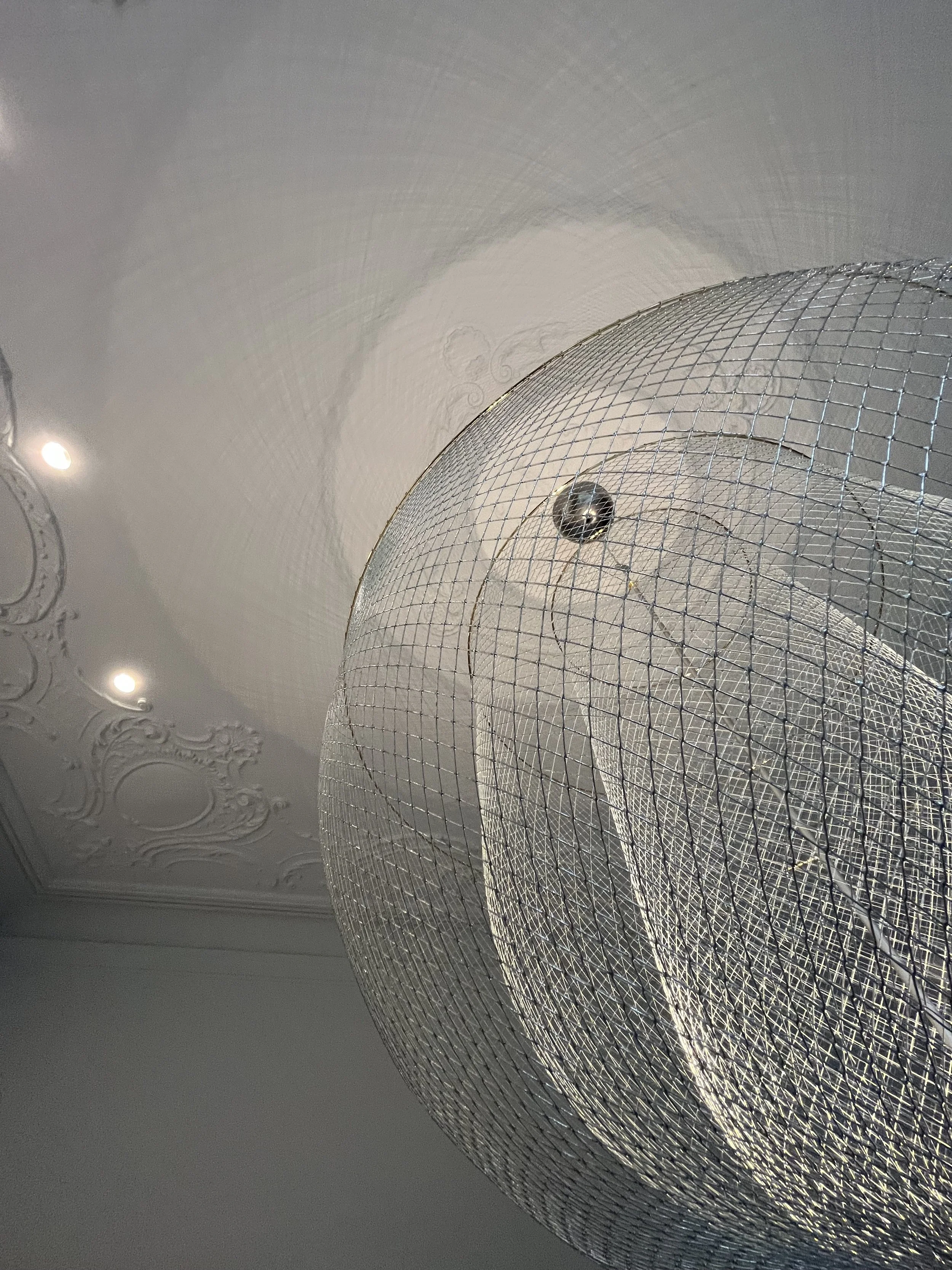 Close-up of a modern chandelier with a wire mesh design, hanging from a decorative ceiling with intricate moldings.