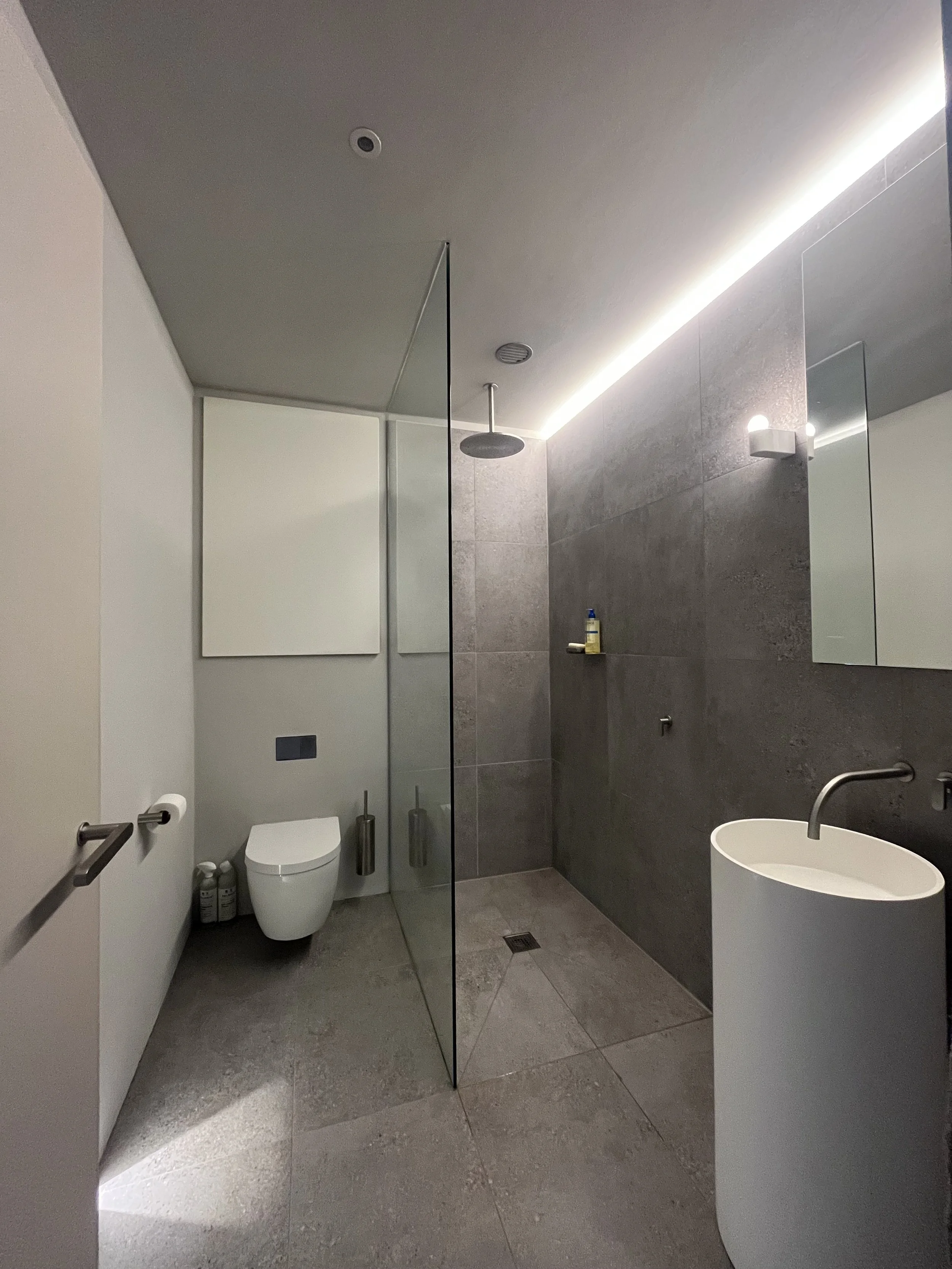 Modern bathroom with gray tiled walk-in shower area, white toilet, white freestanding sink, glass partition, and LED lighting along ceiling.