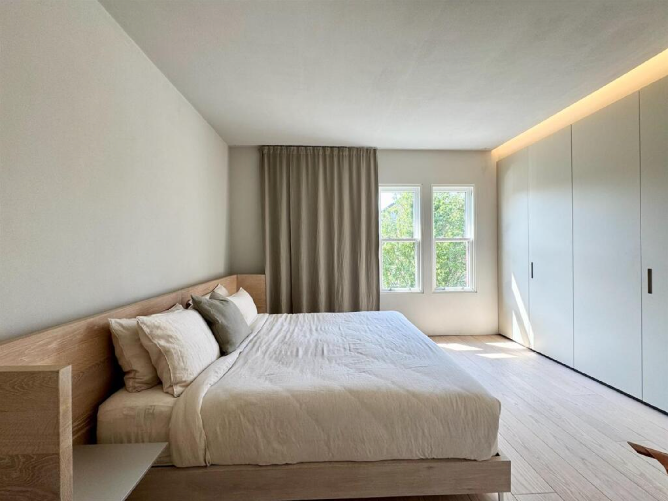 A minimalist bedroom with a large bed, white bedding, and multiple pillows. There are two windows with green trees visible outside, beige curtains, a wooden headboard, and a built-in wardrobe with sliding doors on the right.