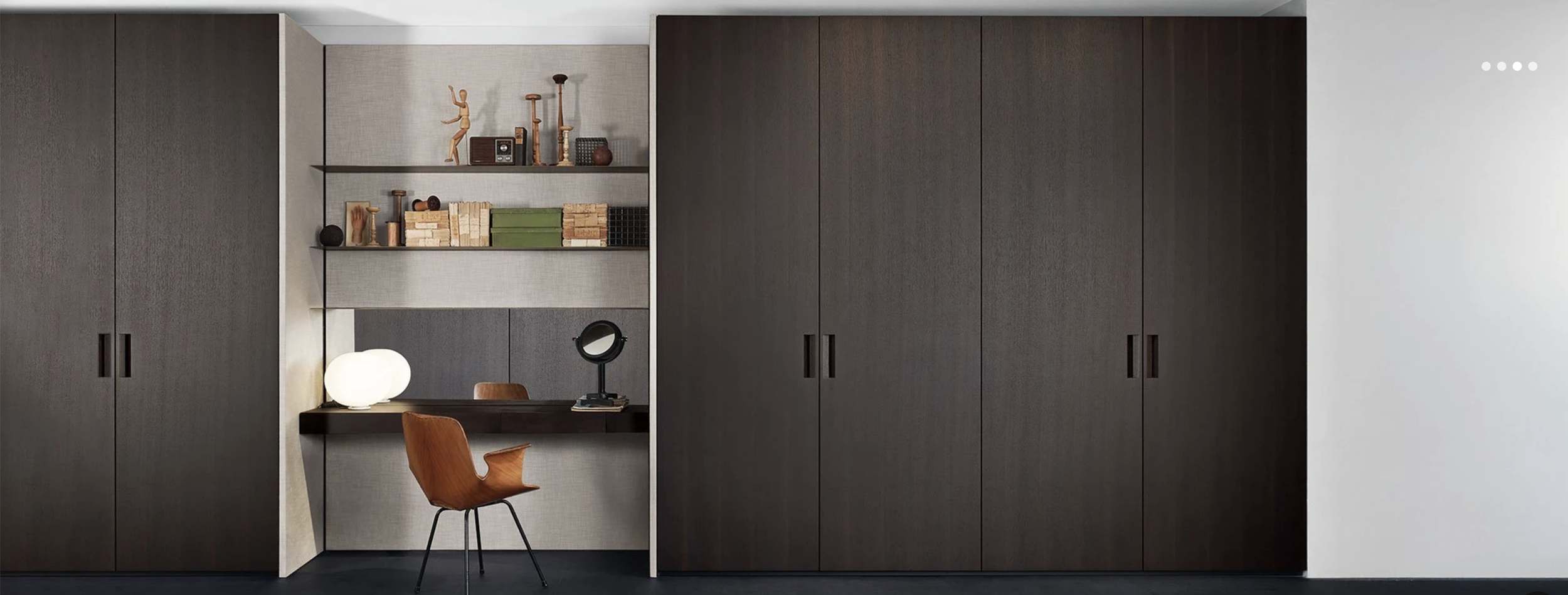 Modern home office with dark wood cabinets, open shelves with decor and books, desk with lamp and mirror, and a wooden chair.