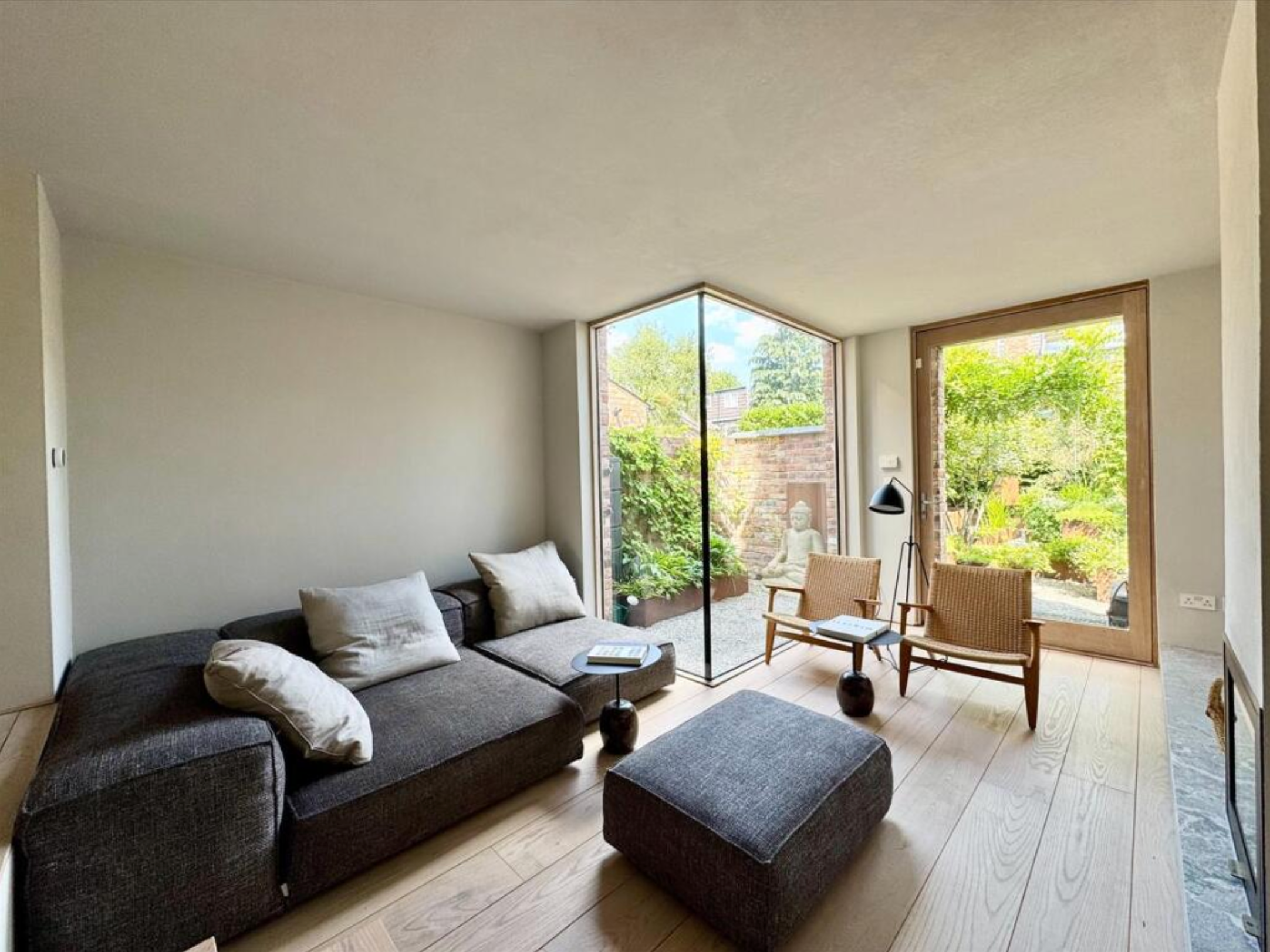 Living room with a large window showing a garden, dark sofa with white pillows, two wooden armchairs, a standing lamp, and a coffee table.