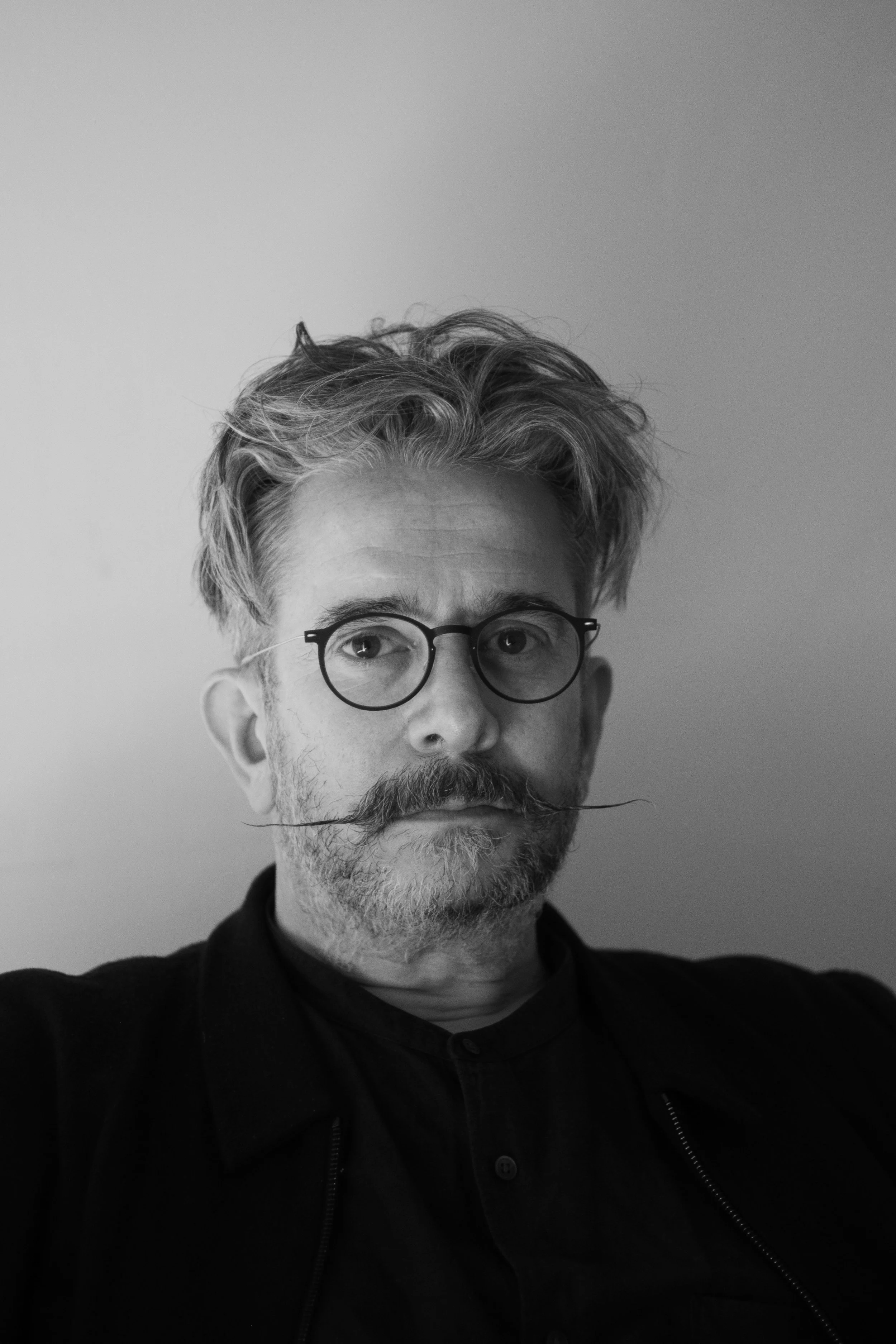 A black and white portrait of a man with wavy, disheveled hair, round glasses, a mustache, and a beard, wearing a dark jacket and shirt against a plain background.