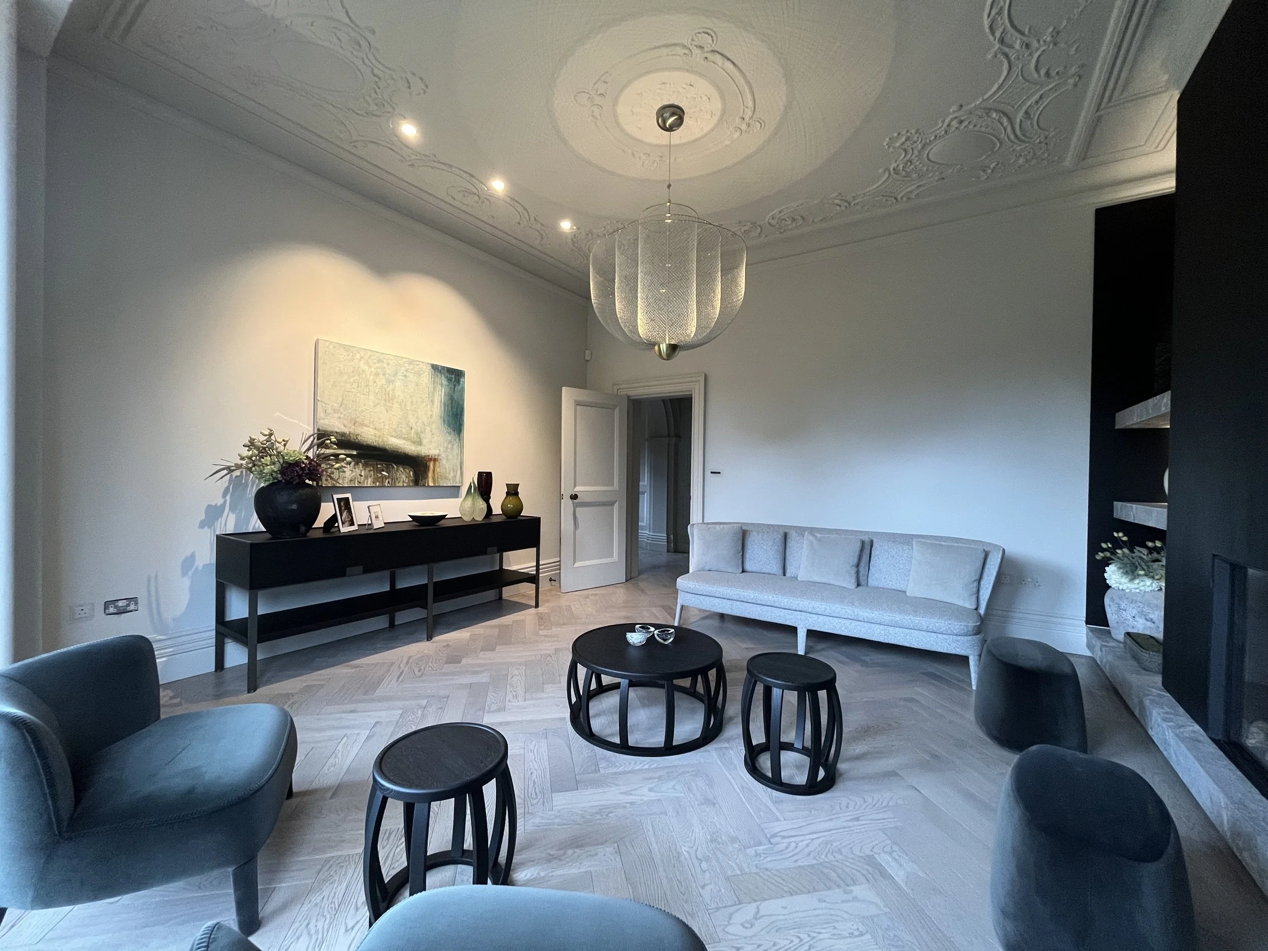 Elegant living room with a high ornate ceiling, a white sofa, gray chairs, a circular black coffee table, and a black sideboard decorated with vases, photographs, and a painting, illuminated by recessed ceiling lights and a decorative hanging chandel