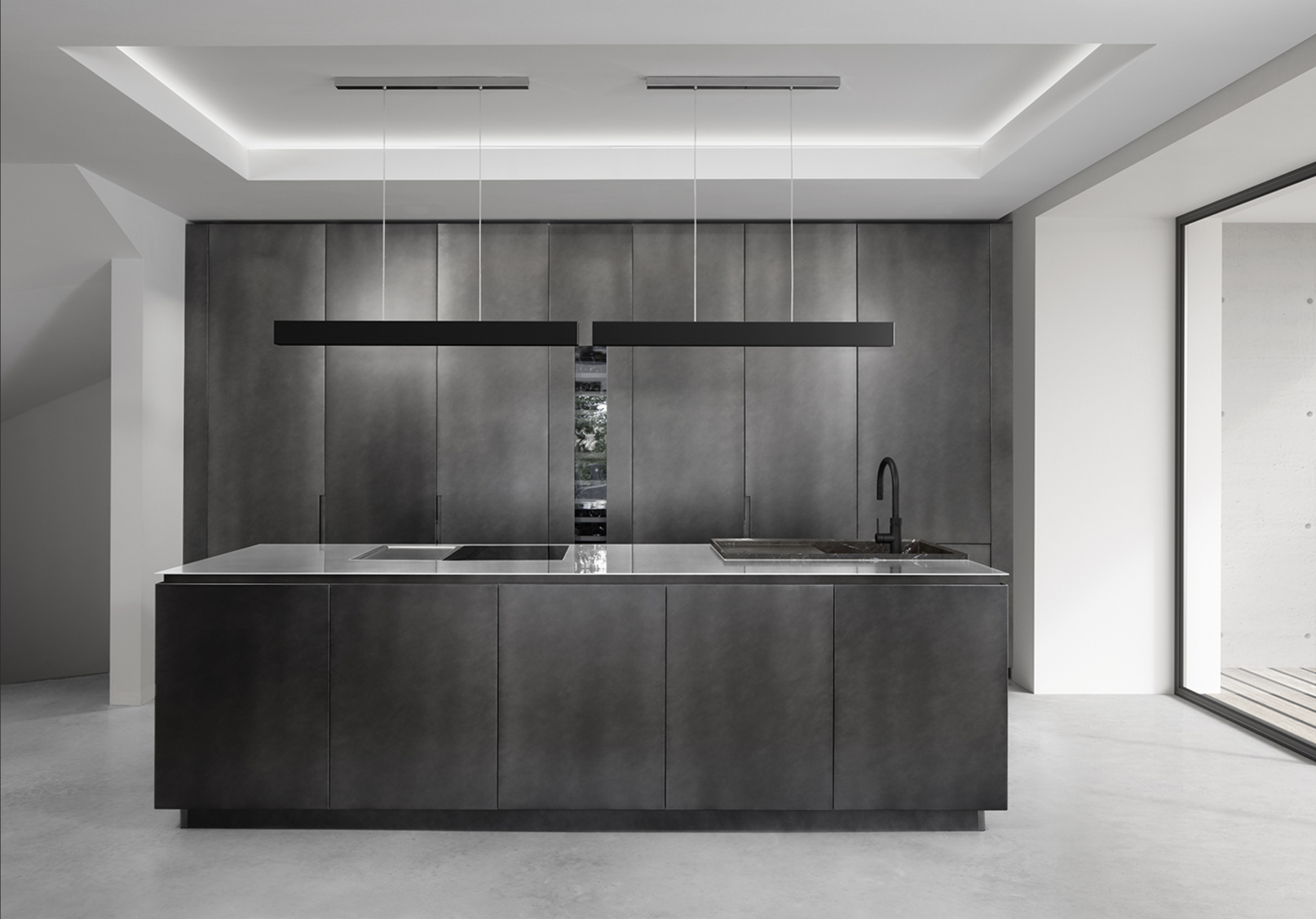 Modern minimalist kitchen with dark cabinetry, a central island with a white countertop, black faucet, gray wall panels, and a large glass sliding door leading outside.