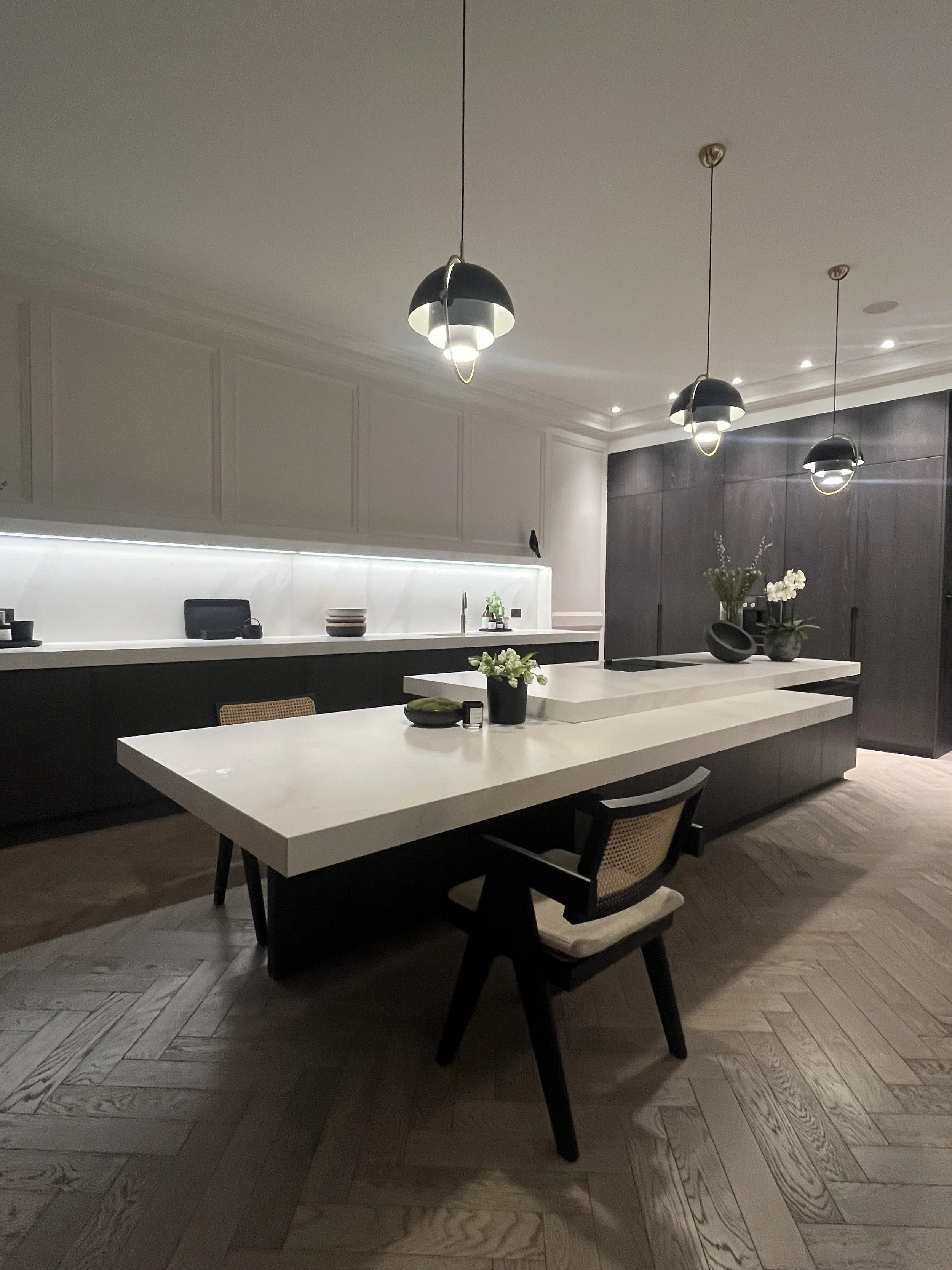 Modern kitchen with white counter and black cabinets, three black pendant lights, and a wooden herringbone floor. Decor includes vases with flowers and tableware.