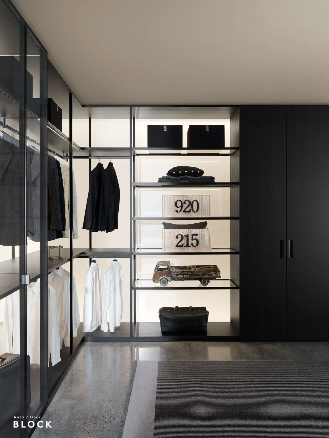 Modern walk-in closet with black shelving and glass doors, holding hanging clothes, storage boxes, decorative objects, and a small black bag.