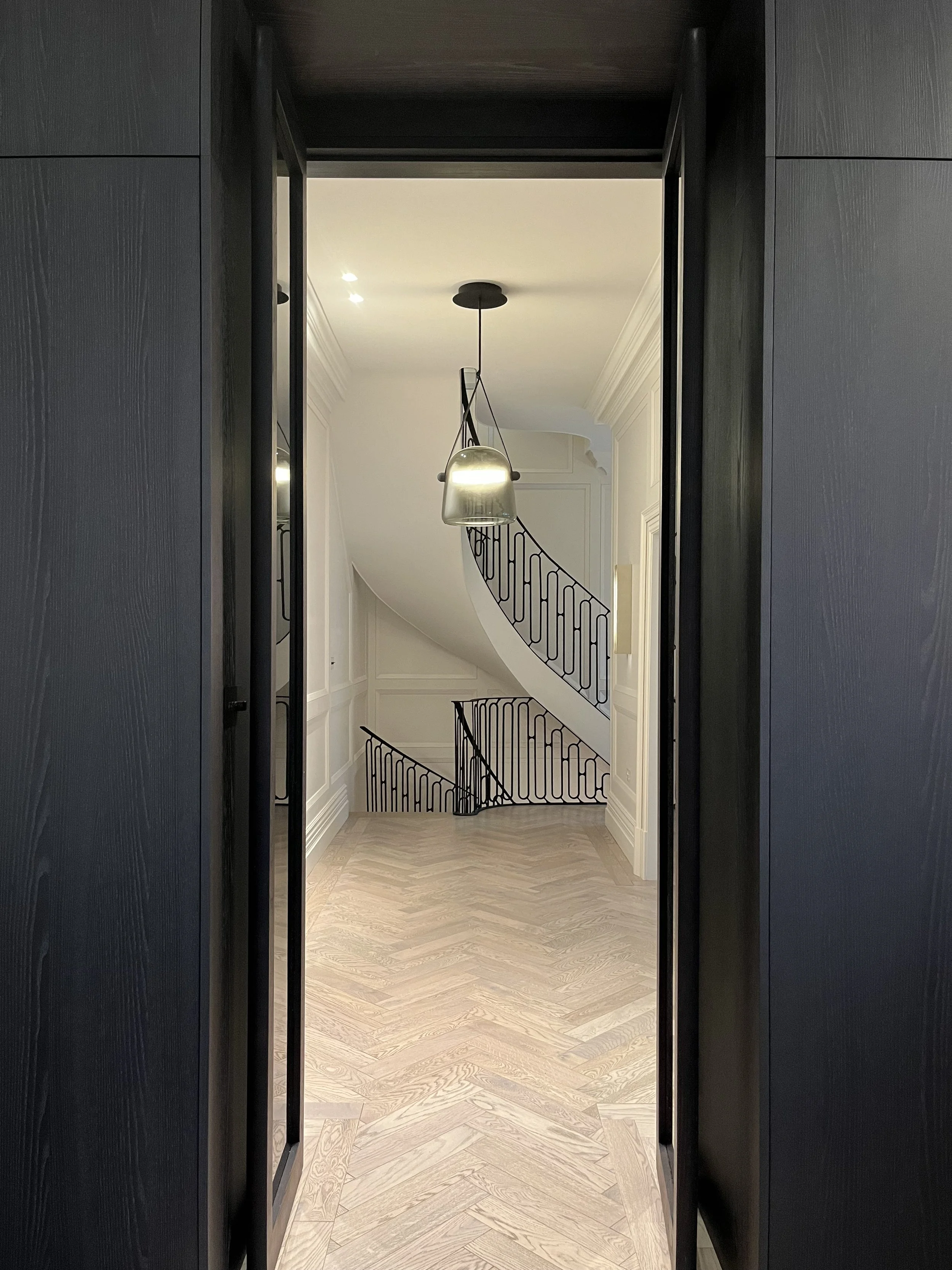 View of a staircase with black metal railing and a modern hanging light fixture, seen through a doorway with dark wood trim, inside a well-lit, elegant home interior.