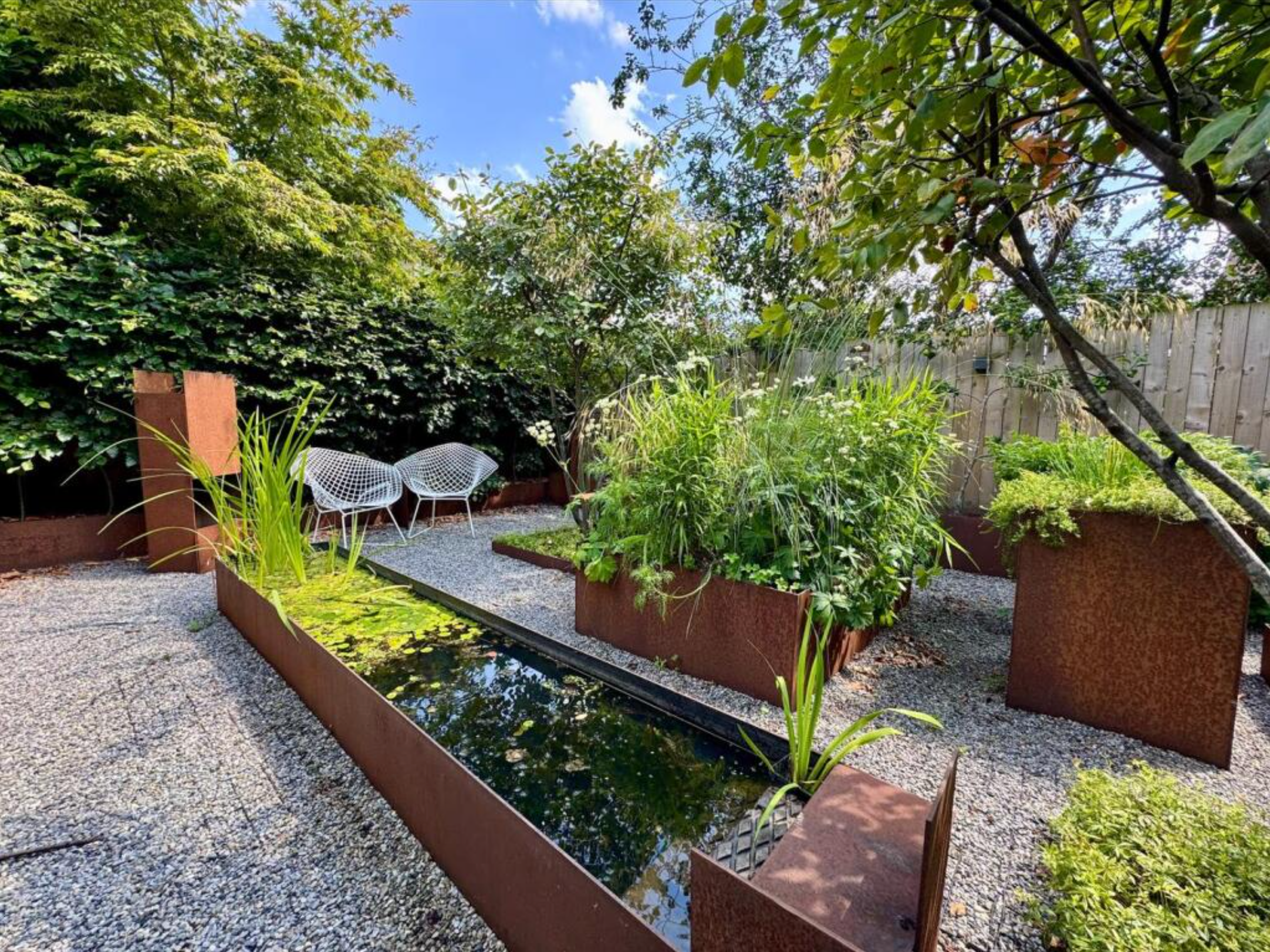 A small garden with raised beds containing various green plants, a narrow water feature with floating water plants, gravel pathways, two white modern chairs, tall trees and a wooden fence, under a bright blue sky with a few clouds.