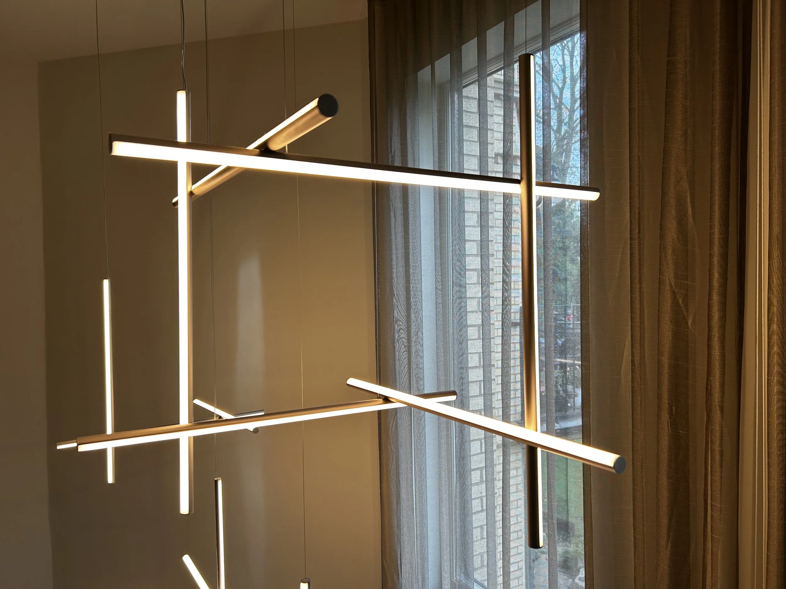 Modern hanging chandelier with horizontal and vertical LED light bars in front of a window with beige curtains and a brick building outside.