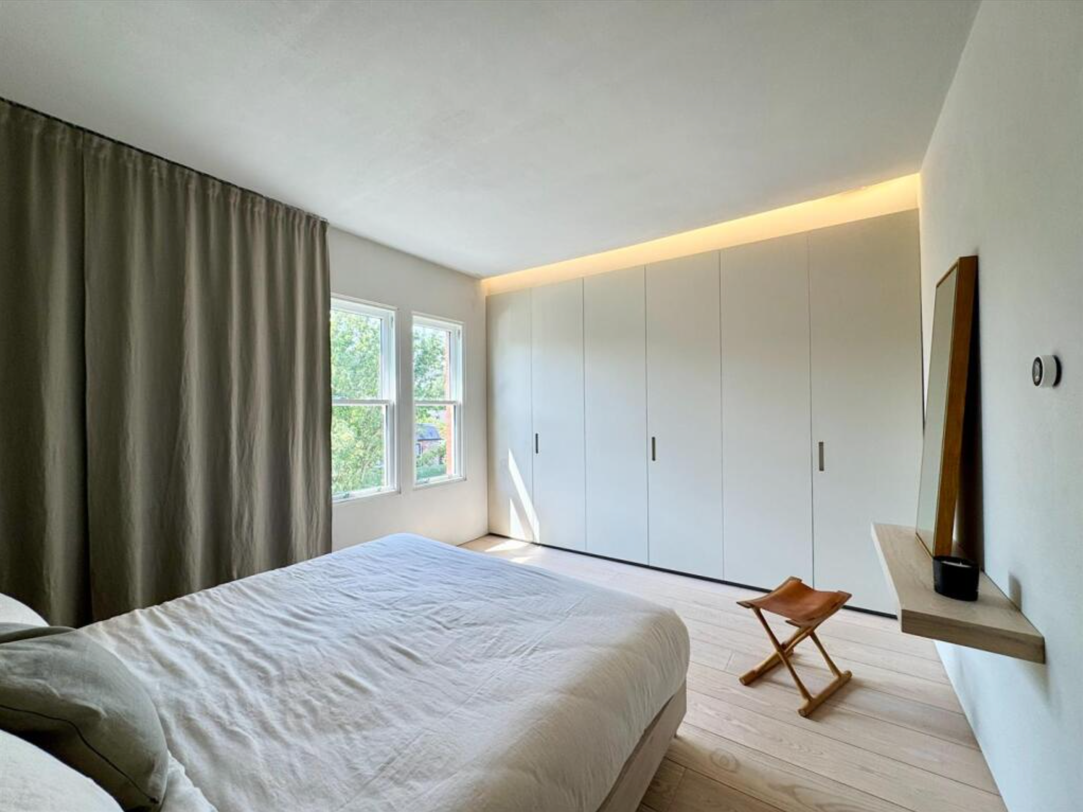 Minimalist bedroom with large bed, beige bedding, window with green trees outside, built-in white wardrobe, narrow wooden stool, and a wall-mounted shelf with a mirror and black speaker.