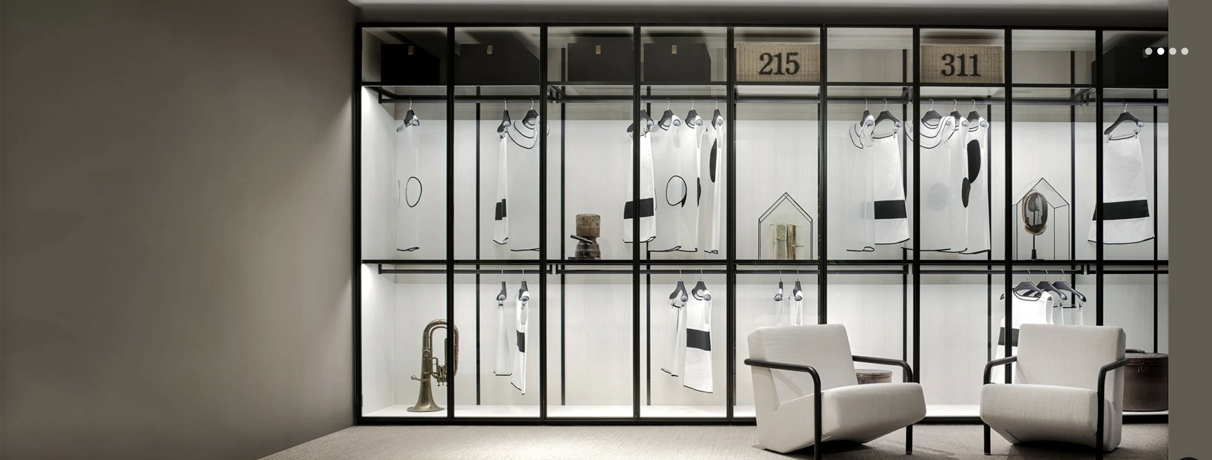 Display case with black metal frame and glass shelves showcasing black and white clothing and accessories, including two white chairs in front.