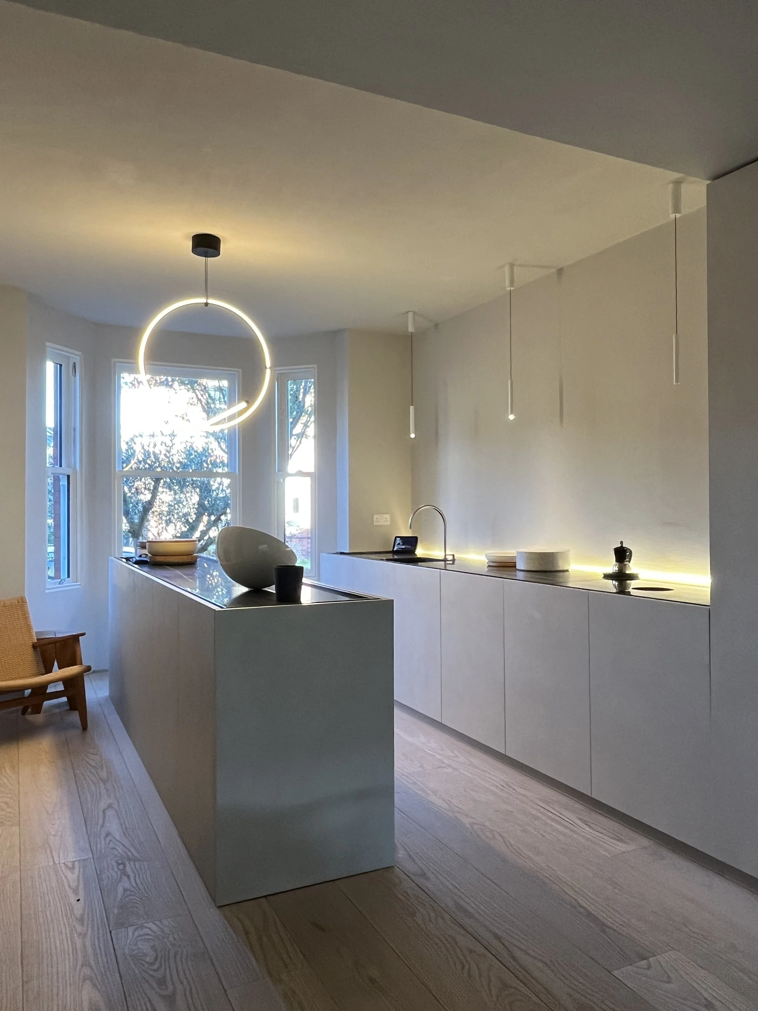 Modern minimalist kitchen featuring white cabinets, a large island with a black countertop, a unique circular pendant light, and three small pendant lights above the counter, with natural light coming through three large windows.