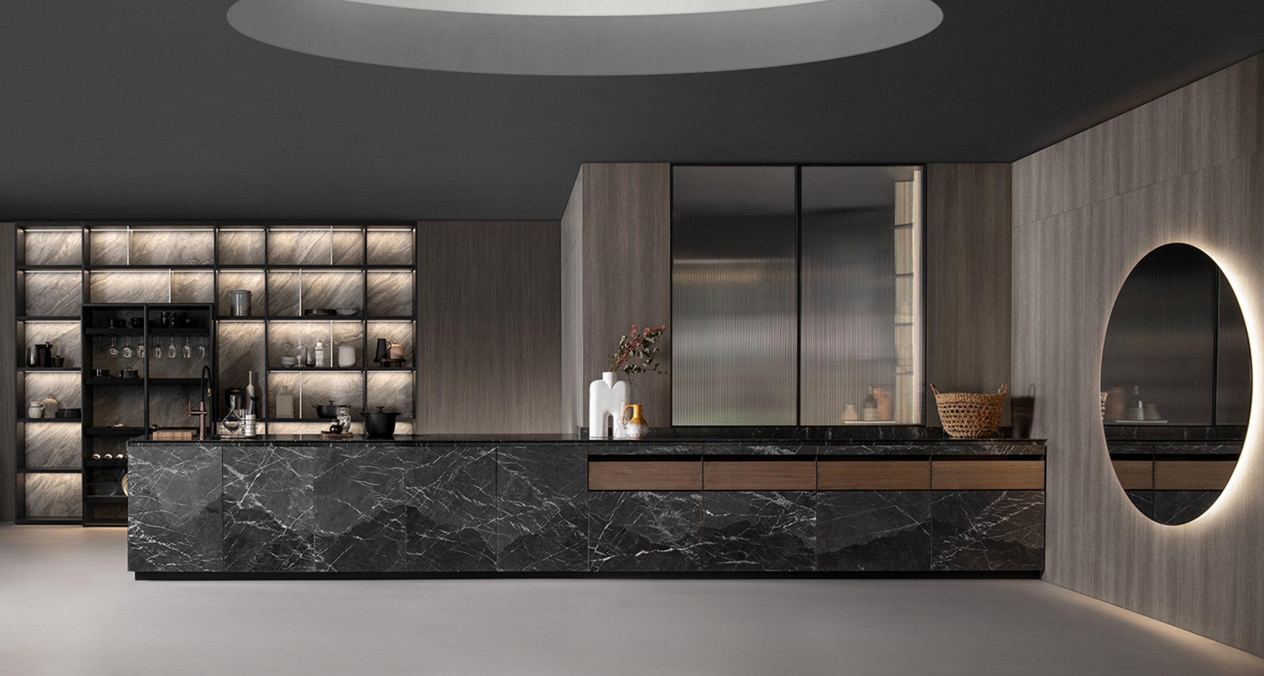Modern kitchen with black marble island, light wood cabinets, open shelving with dishes and glassware, round illuminated mirror on wall, and dark ceiling.