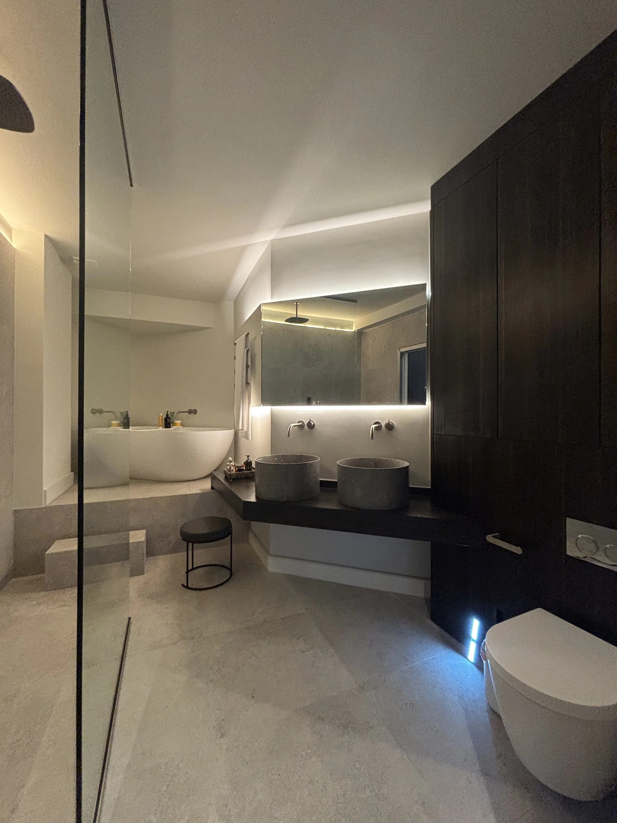 Modern bathroom with stone vessel sinks, large mirror with backlit lighting, dark wooden cabinets, a bathtub, and minimalist decor.