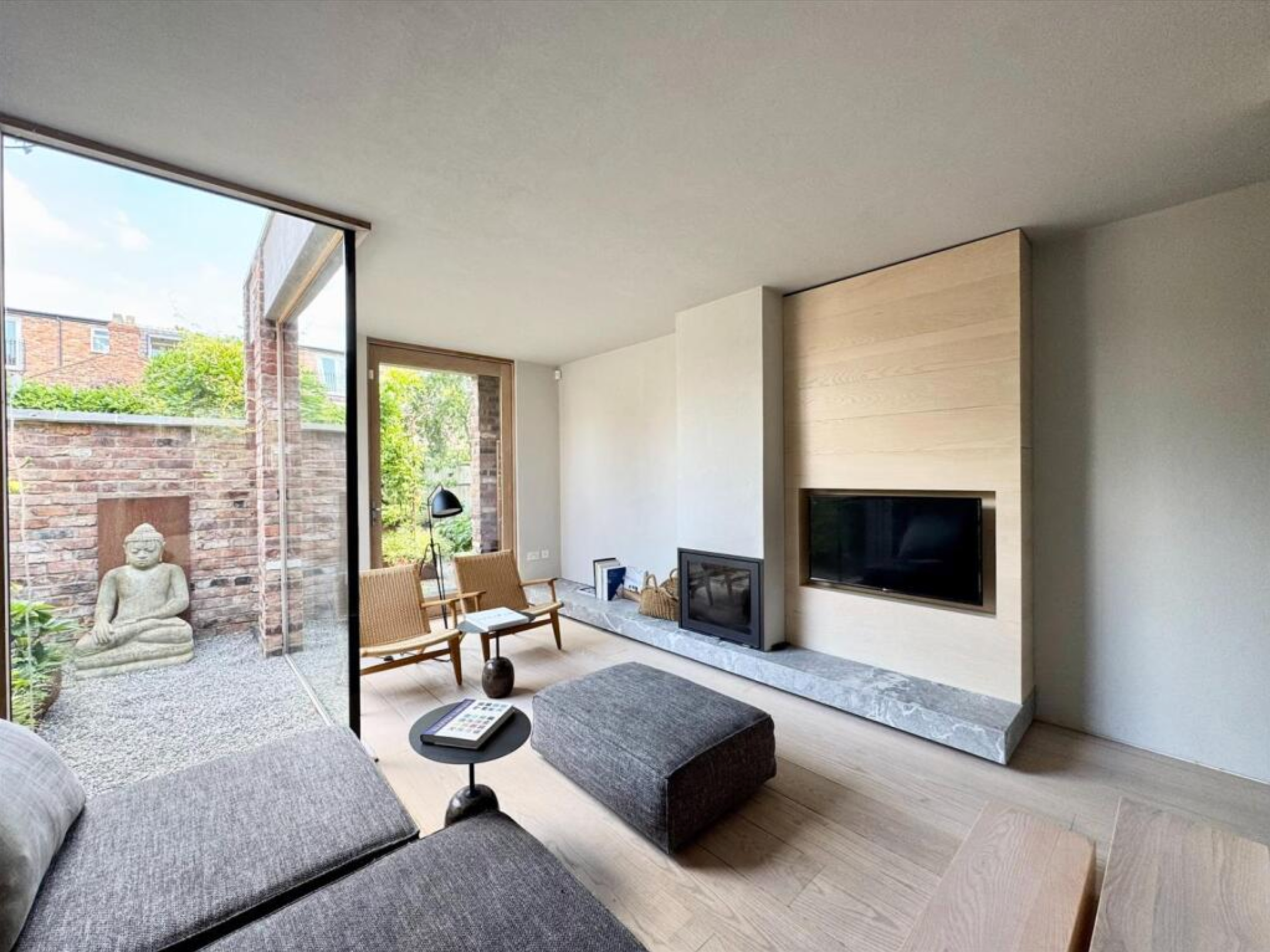 Living room with a gray sofa, a black ottoman, a round black side table with a phone, two wooden armchairs with cushions, a fireplace with a marble base, mounted TV on wood-paneled wall, large glass doors leading to outdoor area with Buddha statue an