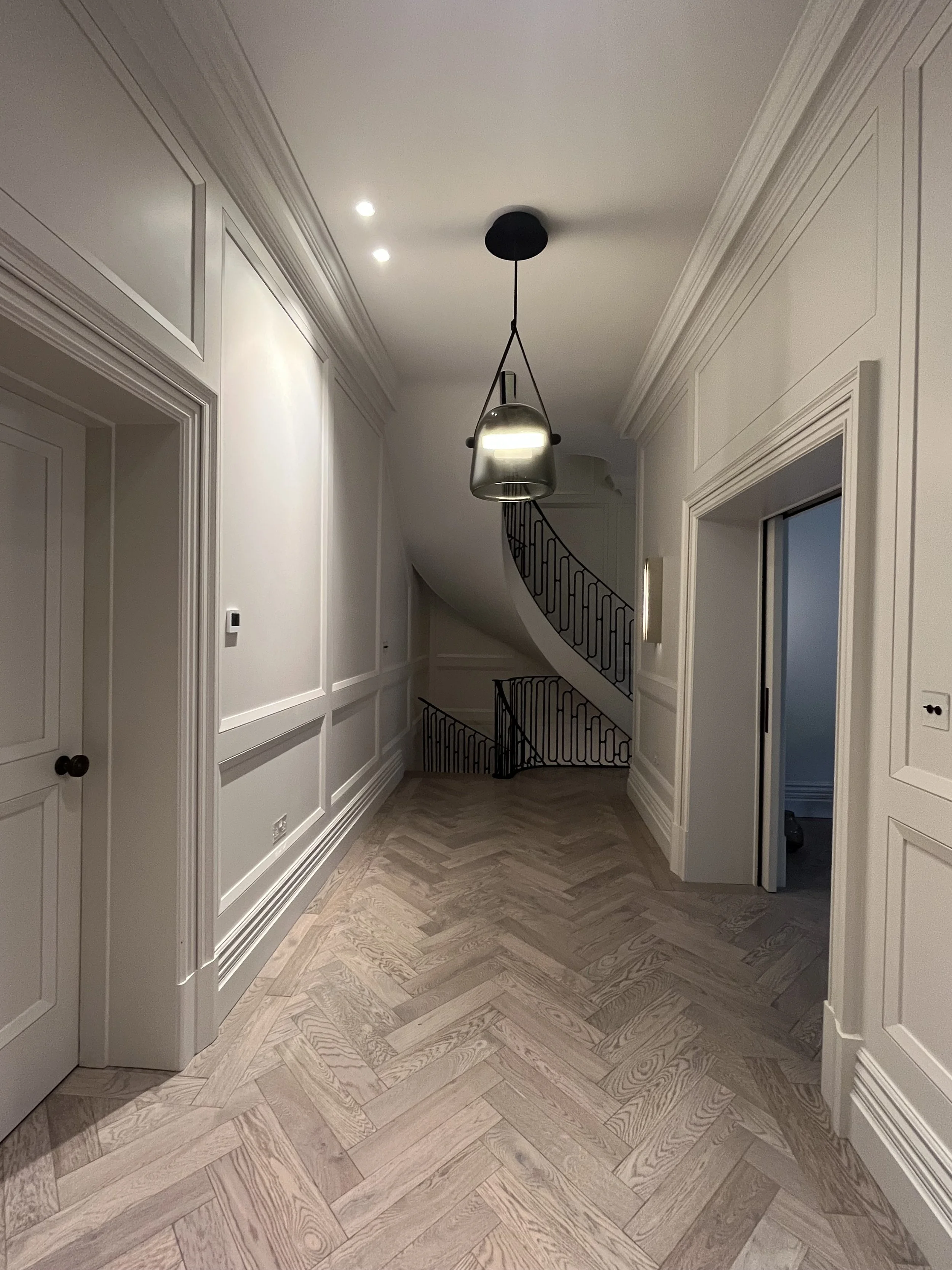 A hallway with wood herringbone patterned flooring, white paneled walls, a black curved staircase with black railing, a ceiling light fixture, and an open door on the right leading to a room. Two small ceiling lights are also visible.