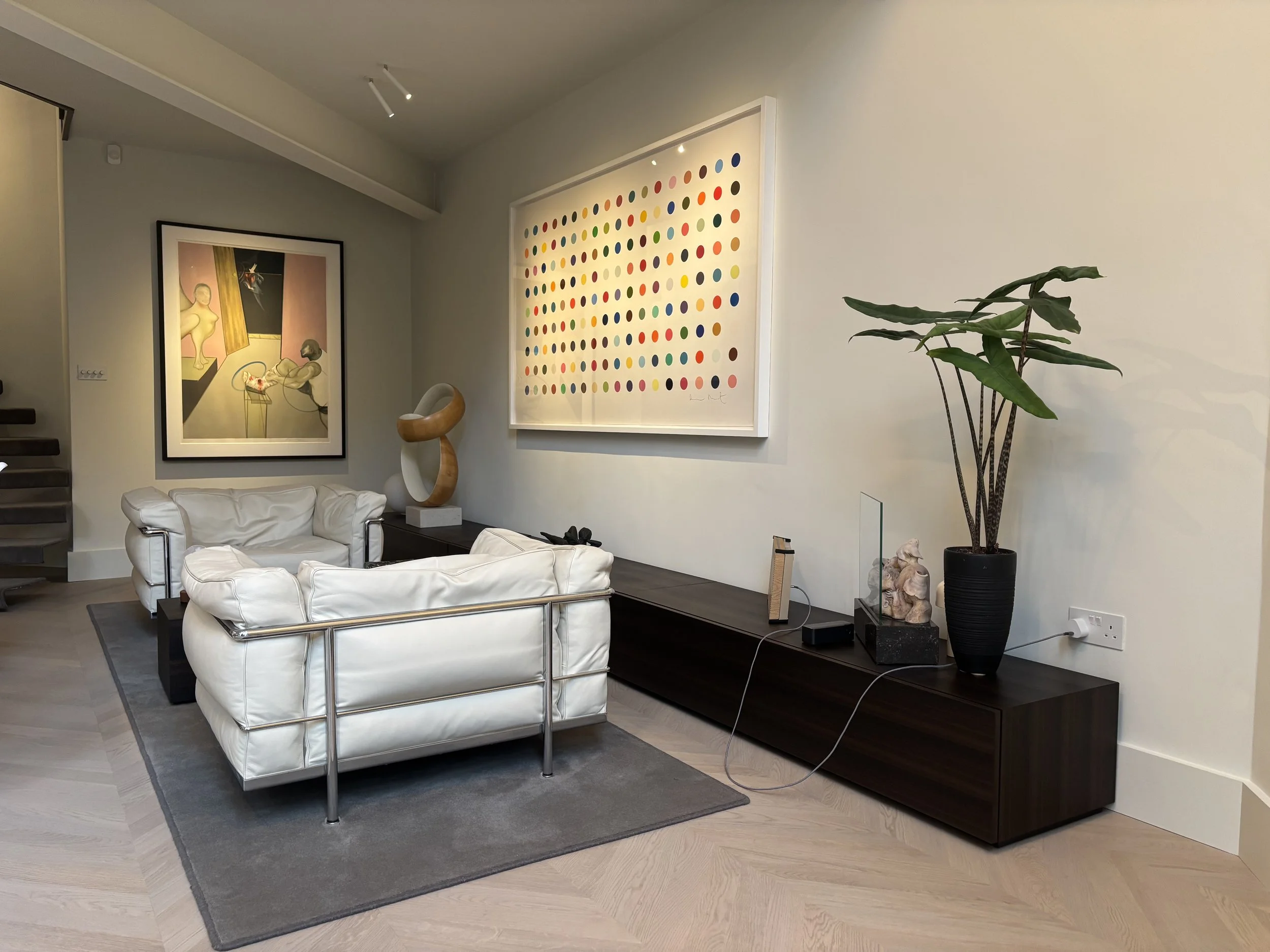Living room with two white leather sofas on a gray rug, dark wooden console with artwork, potted plant, and decorative objects, framed contemporary paintings on the wall, minimalistic decor.
