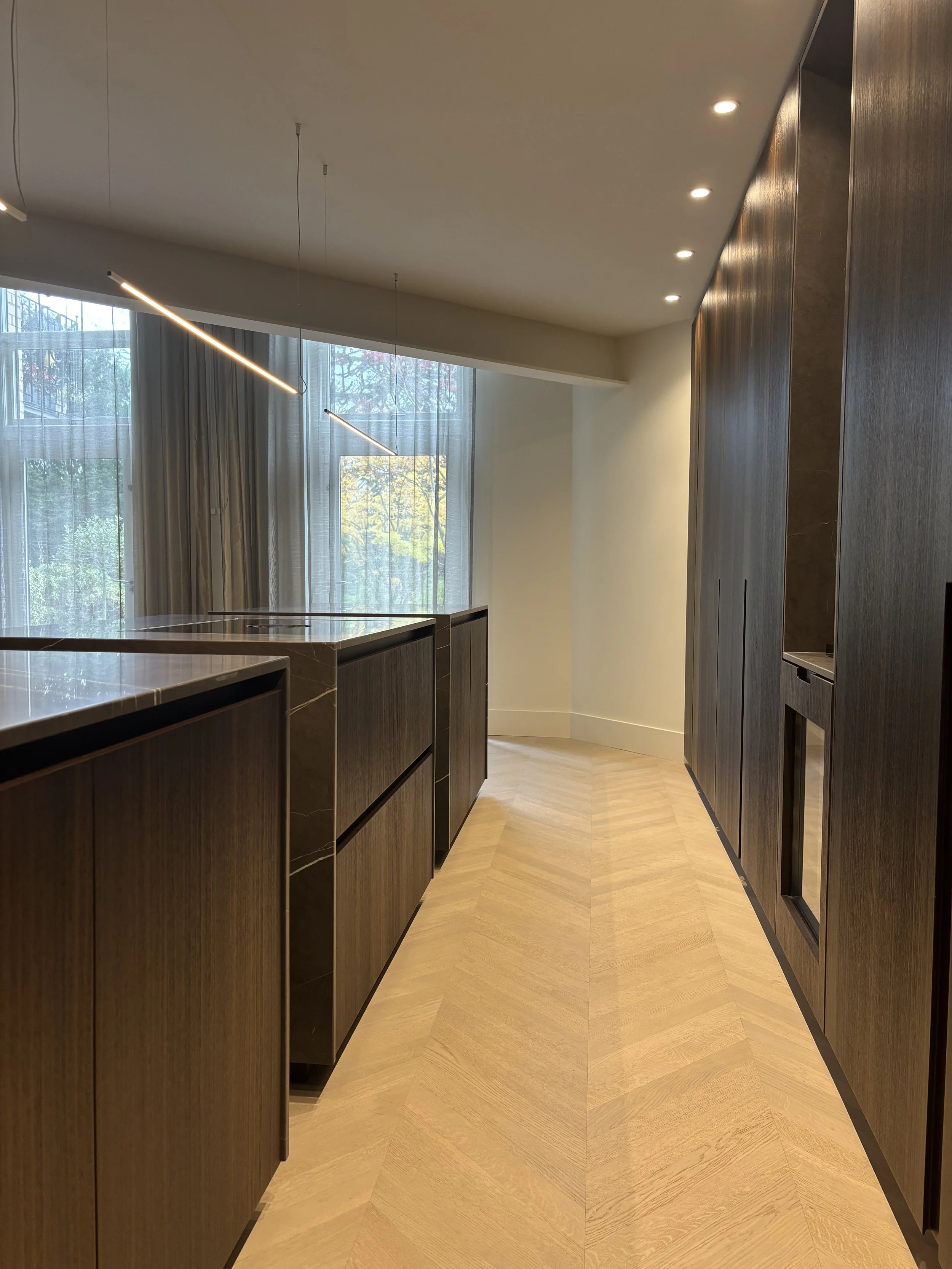 Modern kitchen with dark wood cabinets, light wood chevron-patterned flooring, large windows with sheer curtains, and minimalist LED pendant lighting.