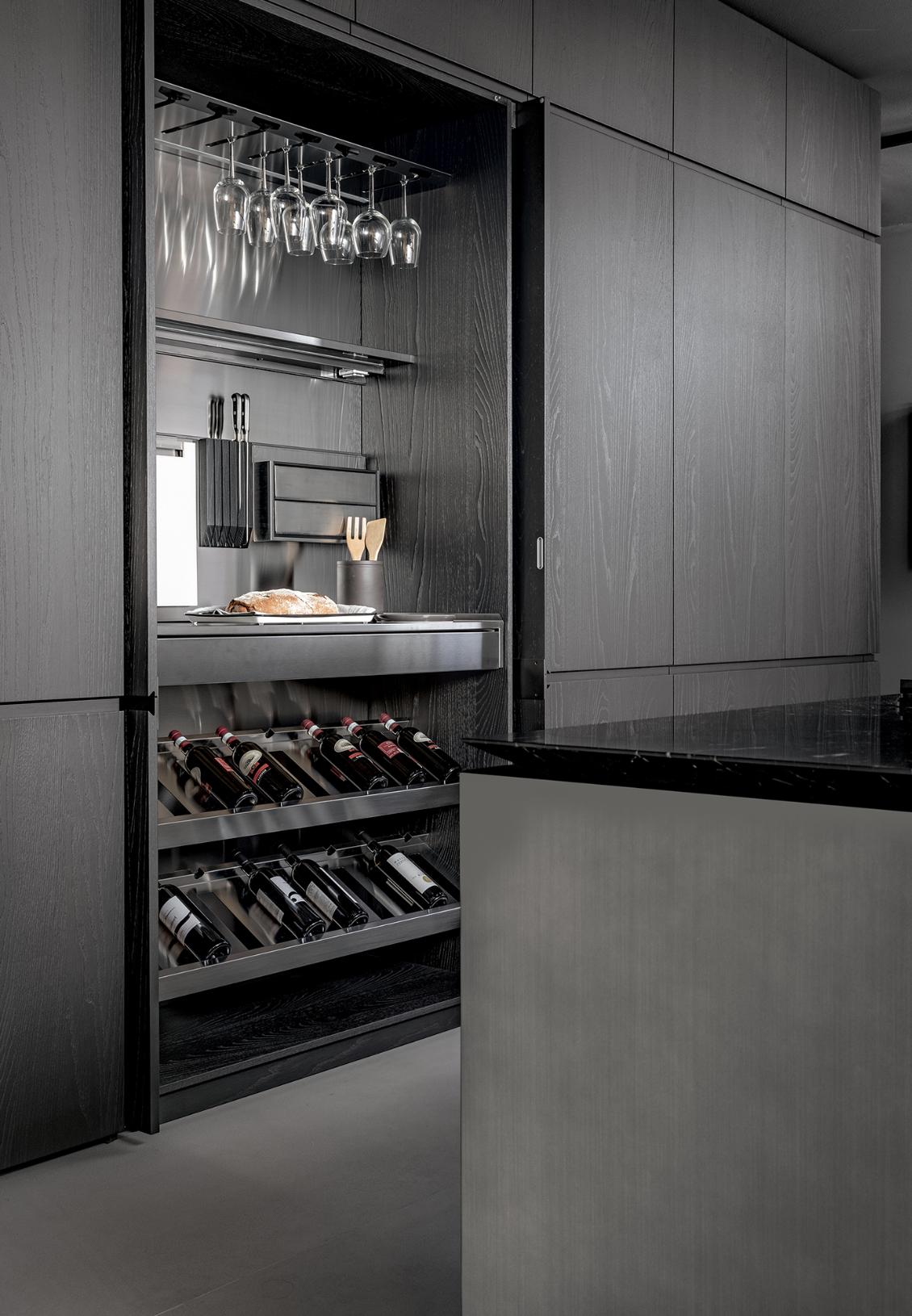 Modern kitchen with built-in wine cooler, wine bottles stored horizontally, hanging wine glasses, and a countertop with a loaf of bread and utensils.