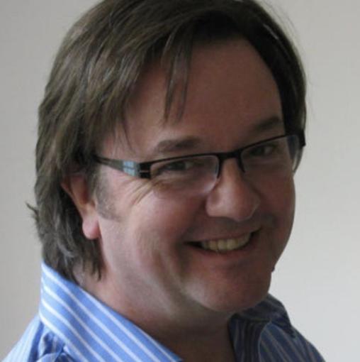 A man with glasses and medium-length brown hair smiling, wearing a blue striped shirt.