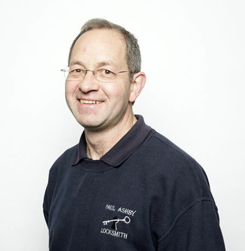 Portrait of a smiling man wearing eyeglasses and a navy blue polo shirt with "Paul Ashby Locksmith" logo, standing against a plain white background.