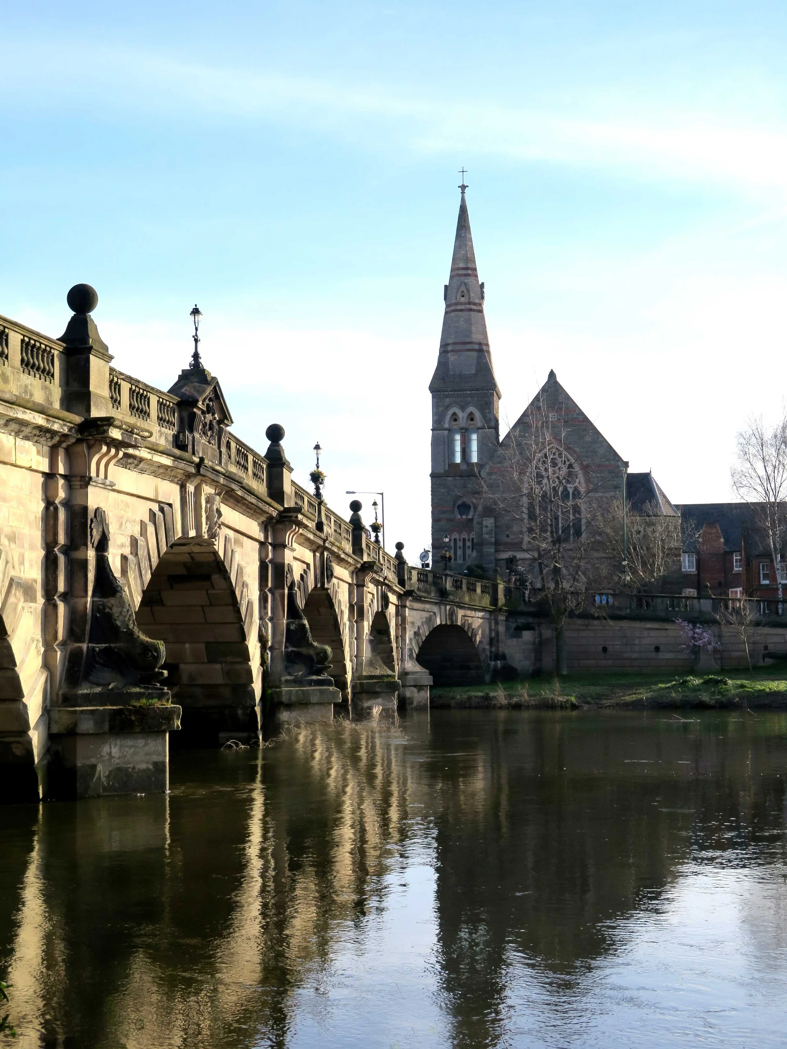 A stone bridge over a river with multiple arches, adjacent to a historical church with a tall steeple, under a clear sky.