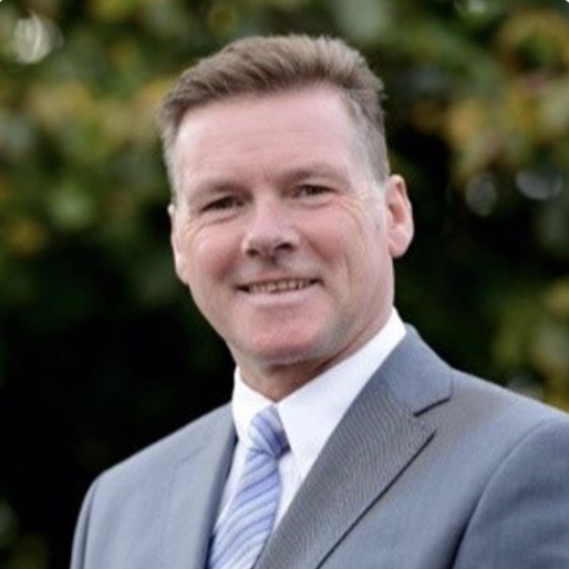 A man in a light gray suit with a white shirt and a light blue tie, standing outdoors with blurred green foliage in the background.