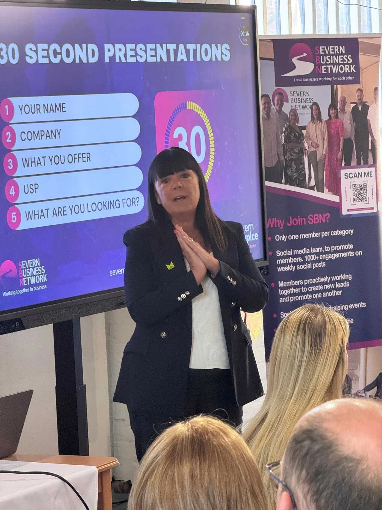 A woman giving a presentation in front of a screen with the title '30 SECOND PRESENTATIONS' and a list that includes 'Your Name,' 'Company,' 'What You Offer,' 'USP,' and 'What Are You Looking For?'. There is also a banner for 'SEVERN BUSINESS NETWORK' next to her, showing a group photo of diverse people and additional information about joining the network.