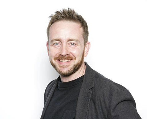 Portrait of a smiling man with short light brown hair and a beard, wearing a black t-shirt and a dark blazer against a white background.
