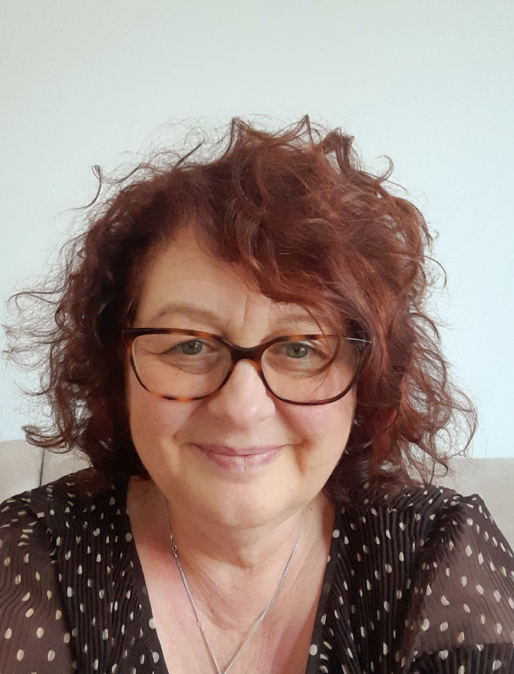 Woman with curly red hair and glasses smiling indoors, wearing a black polka dot top.