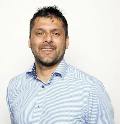 A smiling man with short dark hair and a beard, wearing a light blue striped collared shirt, standing against a plain white background.