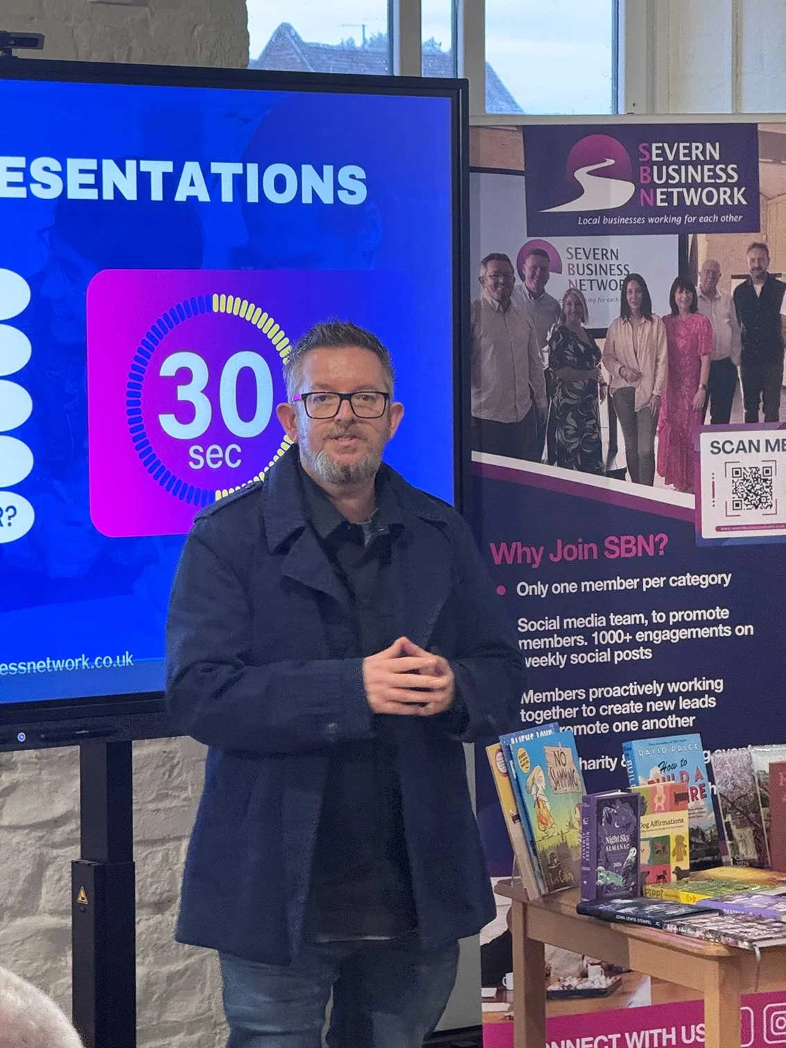 A man with glasses and a beard is standing in front of a presentation screen and a promotional banner for SEVERN BUSINESS NETWORK. The screen displays the word 'PRESENTATIONS' and a 30-second timer. The banner provides reasons to join SBN, such as limited membership categories, social media promotion, member collaboration, and charity support. There is a table with books and a QR code on the banner.