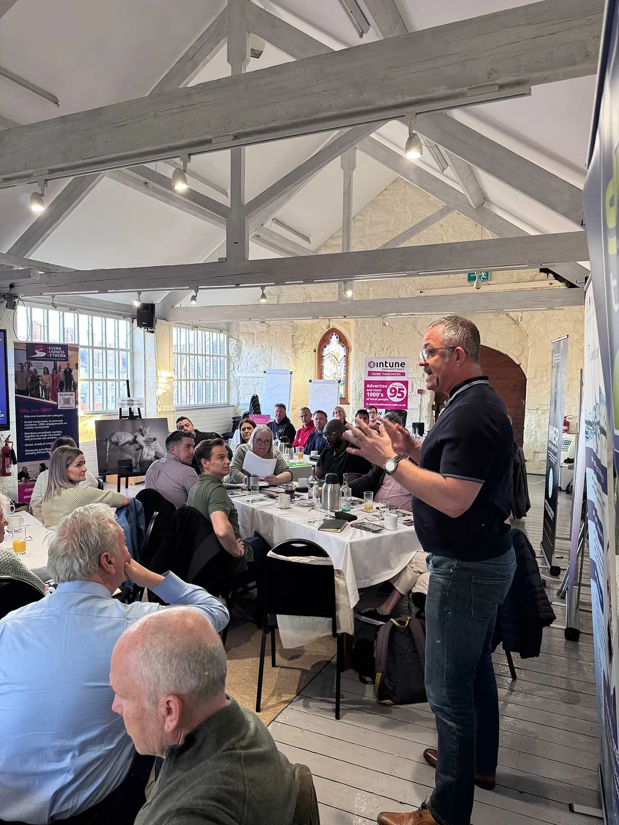 A man standing and speaking in front of a large group seated at tables during a meeting or seminar, with conference materials and a display screen nearby in a well-lit room with high ceilings and wooden beams.