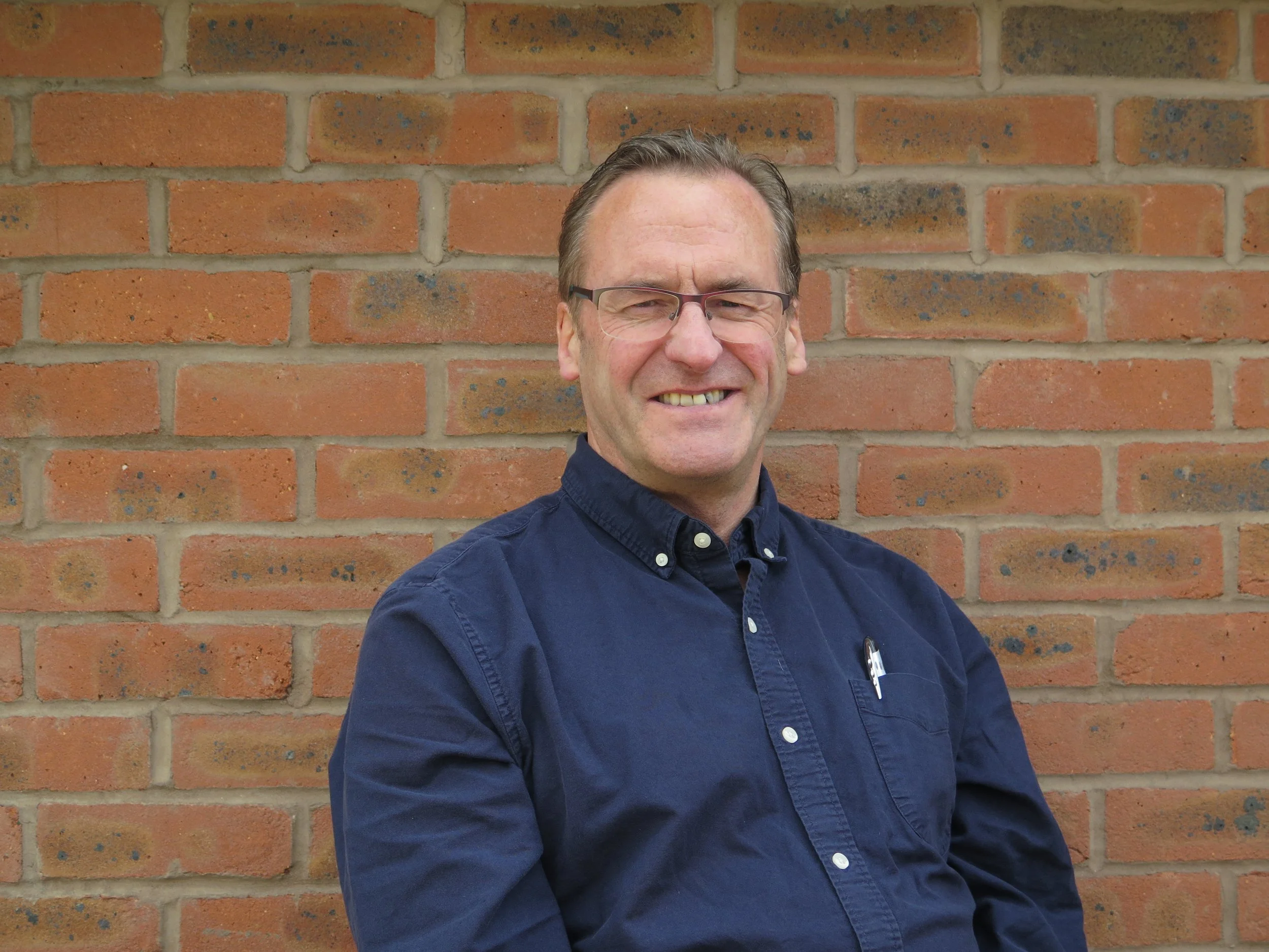 A man with glasses smiling confidently against a brick wall, wearing a navy blue button-up shirt with a pen in his pocket.