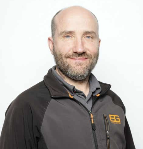 A man with a bald head and beard wearing a black and grey jacket with orange accents and an 'EB' logo, smiling at the camera.